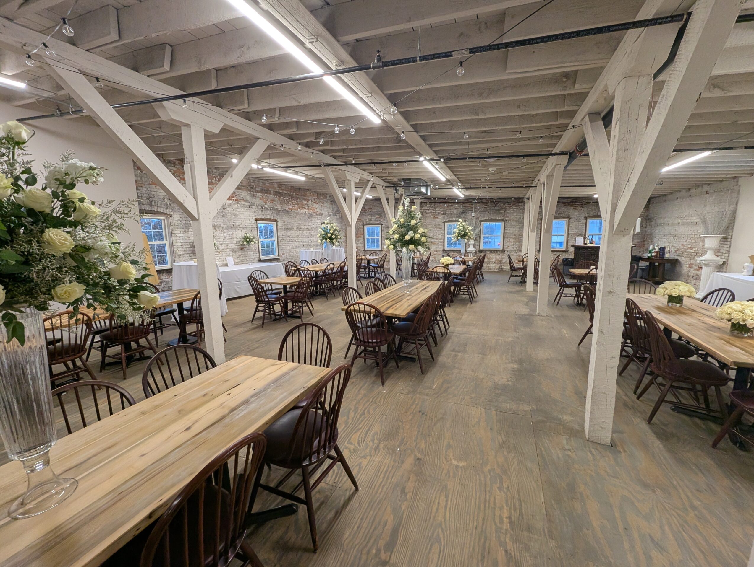 Empty dining area with wooden tables, chairs, white beams, and large windows, decorated with plants and flowers.