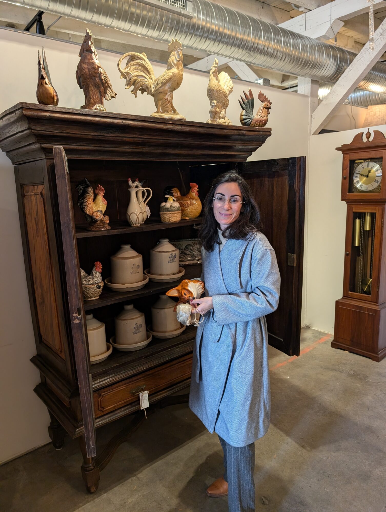Woman in blue coat holding a cup, standing in front of a wooden display cabinet with ceramic and porcelain items.