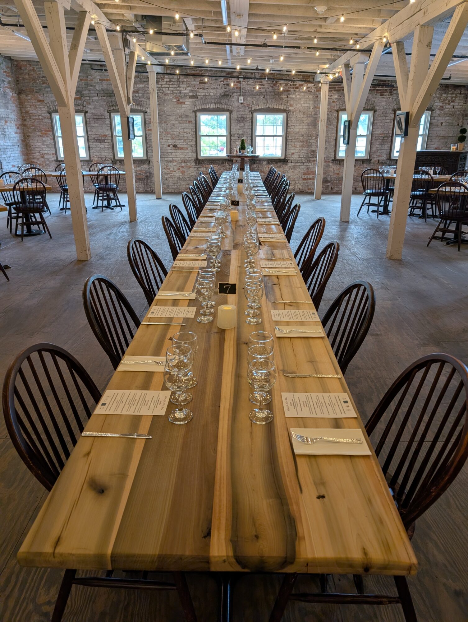 Long wooden table set with glasses, plates, and utensils in a spacious, rustic event hall with brick walls and string lights.