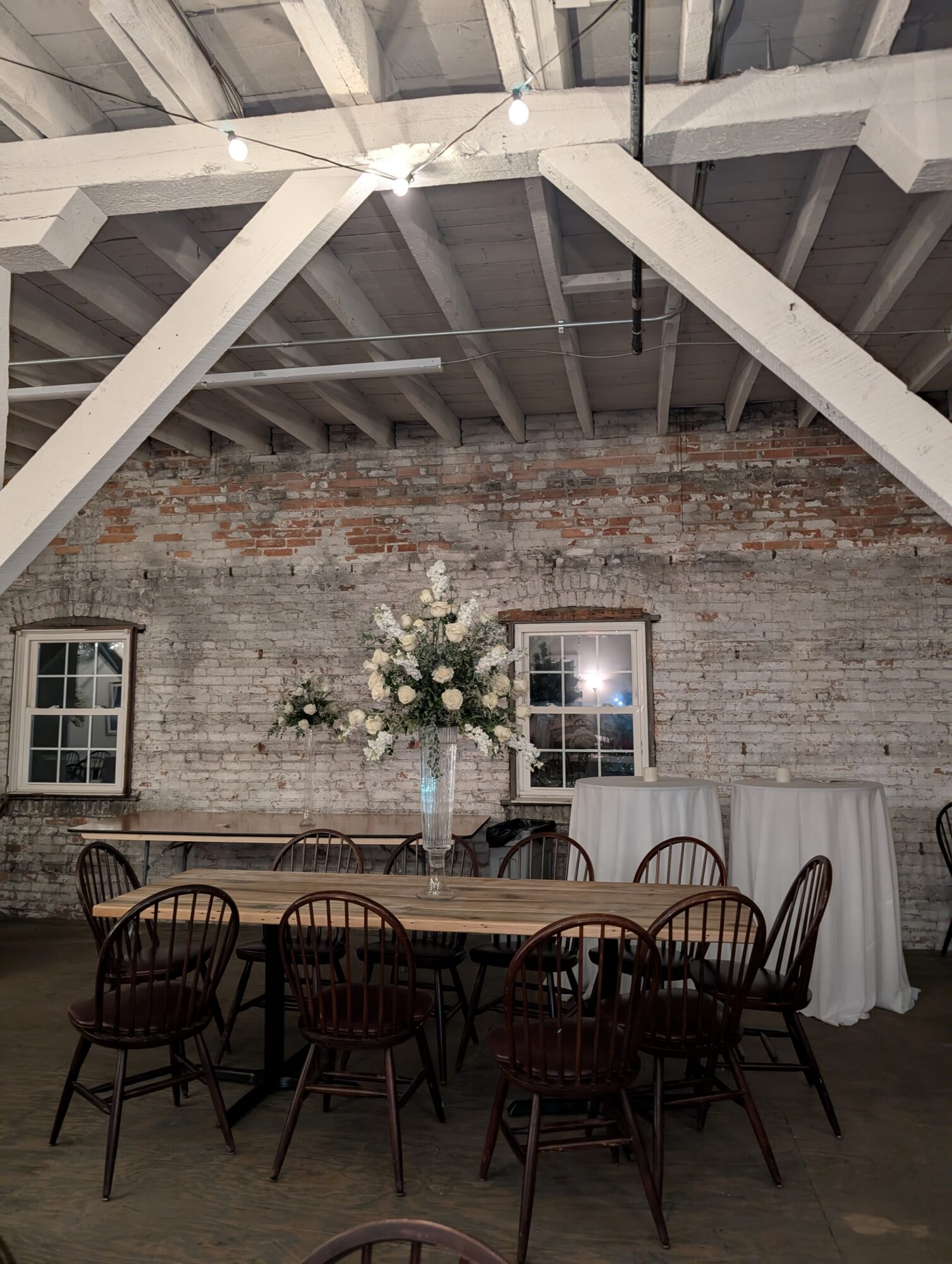 Indoor event space with a round table, chairs, and a floral centerpiece against a brick wall with windows.