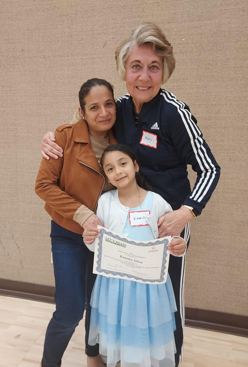 Three people, two women and a girl, standing together indoors, smiling, holding a certificate, beige wall background.