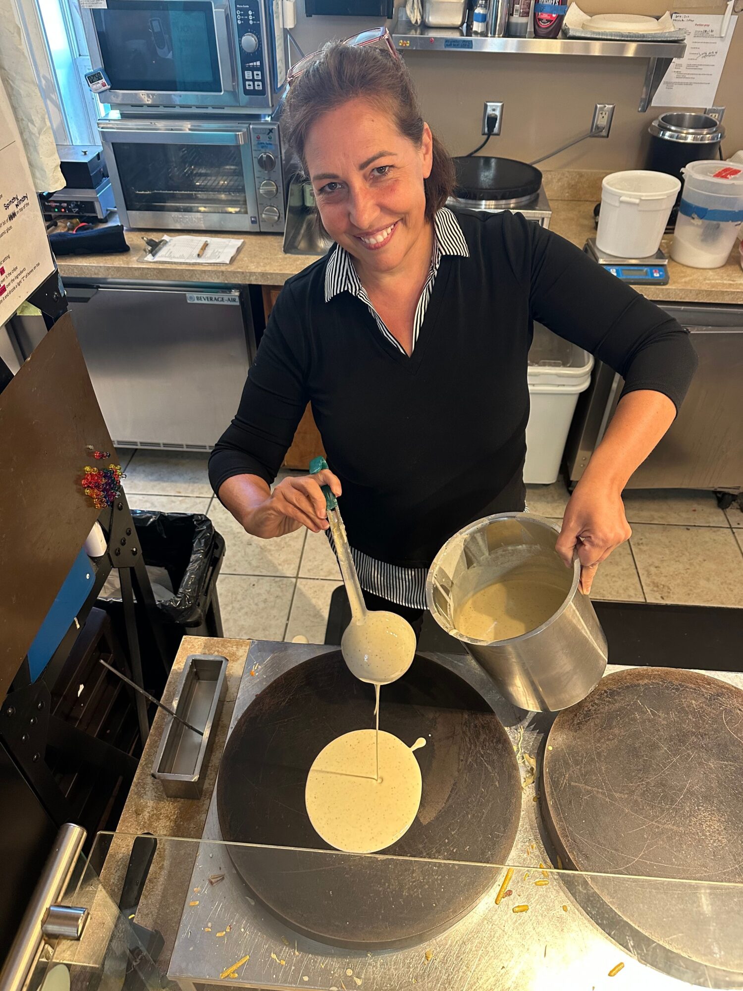 Woman pouring pancake batter onto a griddle in a kitchen, smiling, with kitchen appliances and supplies in background.