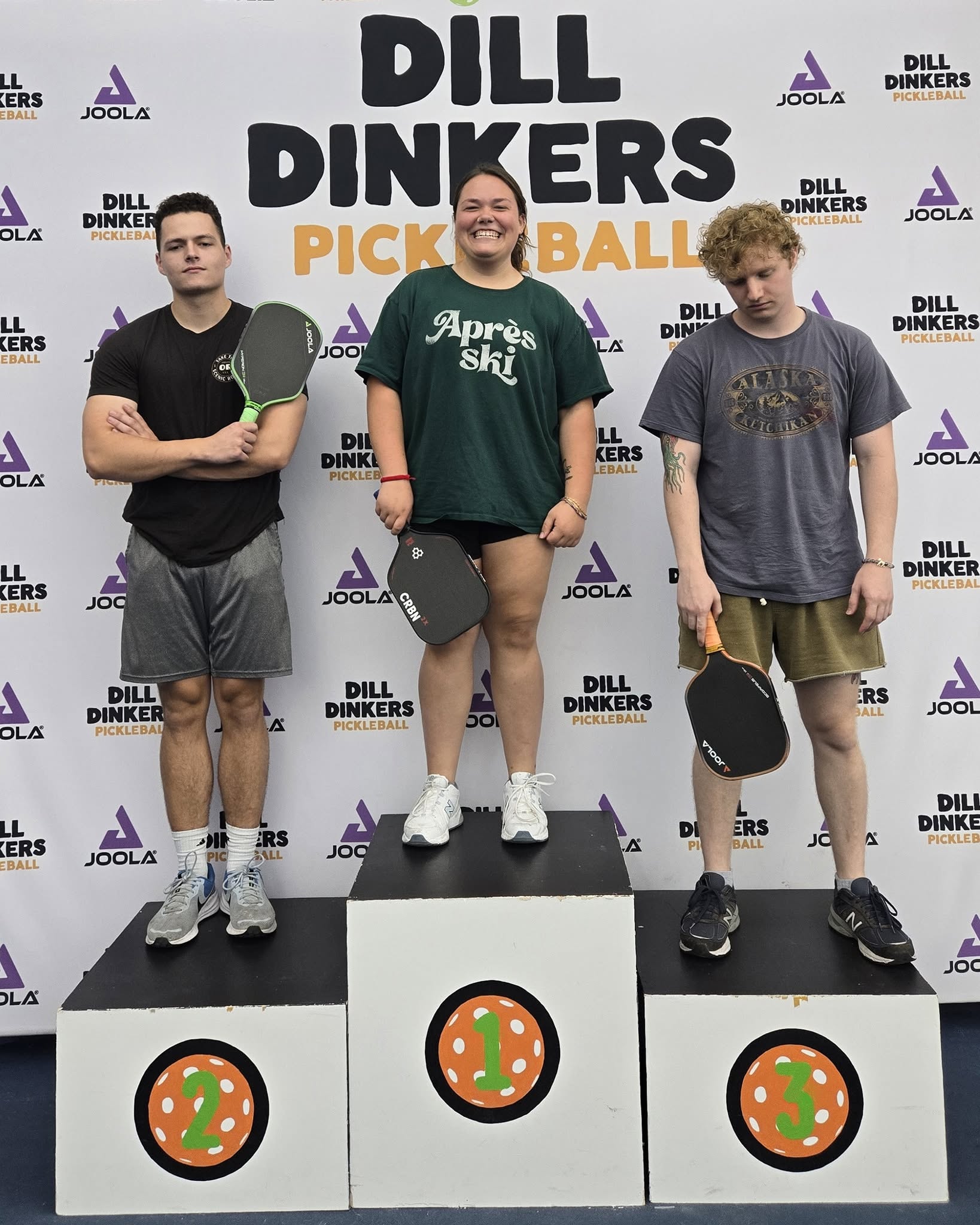 Three young people standing on a winners' podium with a backdrop for Dill Dinkers Pickleball event, holding paddles.