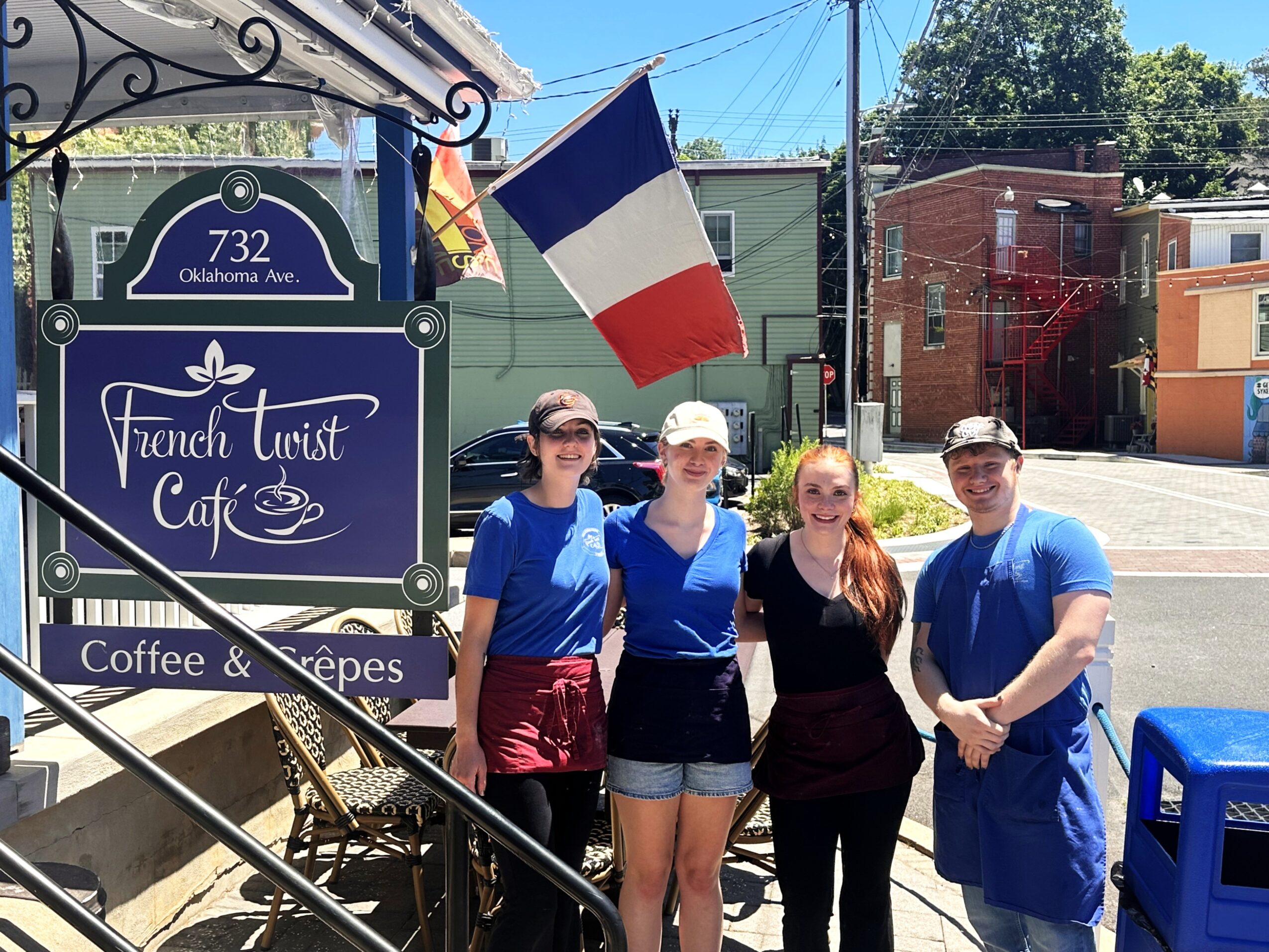 Four people stand outside a cafe with a French flag, smiling, in front of a sign for French Tourist Cafe.