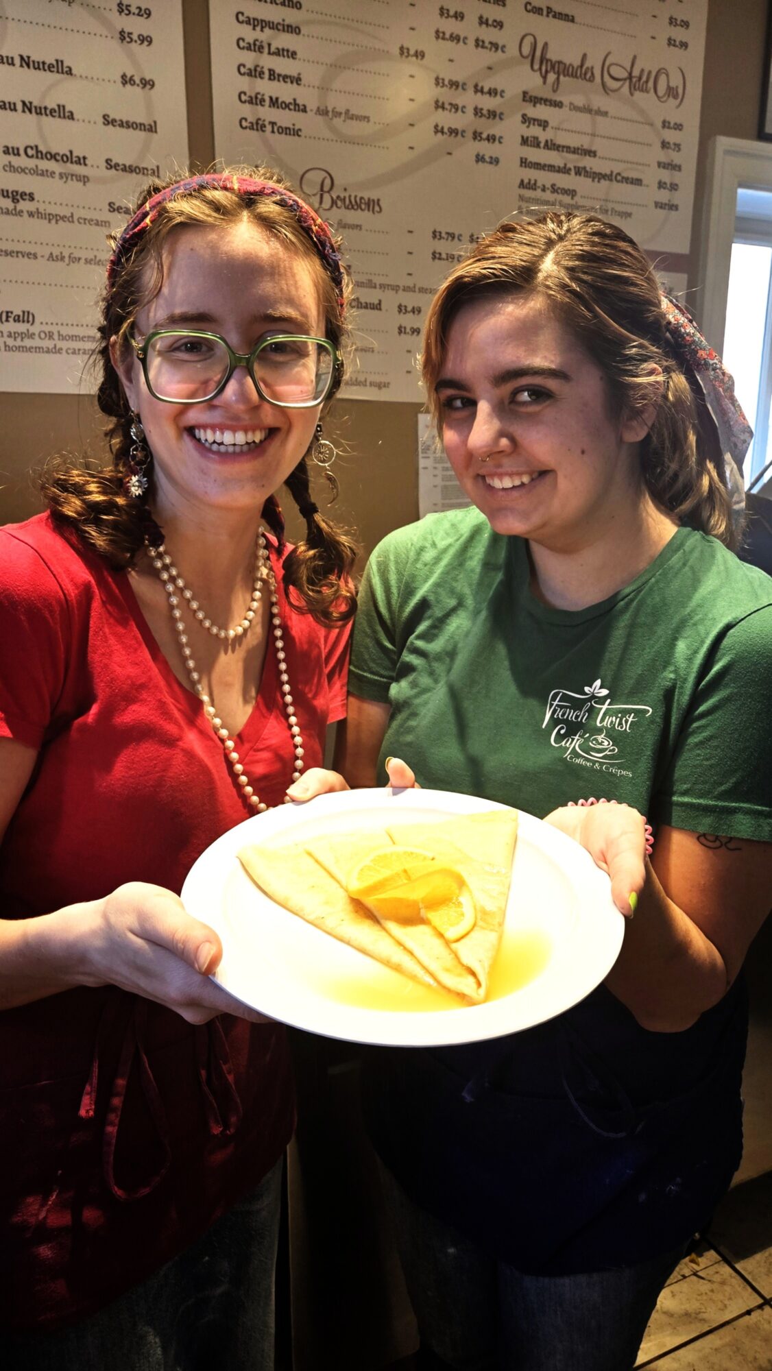 Two women smiling, one holding a plate with a slice of cake, in a cafe setting.