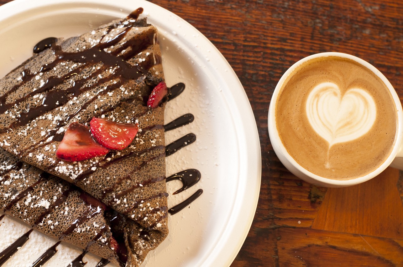 Plate of folded crepes with chocolate drizzle and strawberries, next to a cup of coffee with heart-shaped foam, on wooden table.