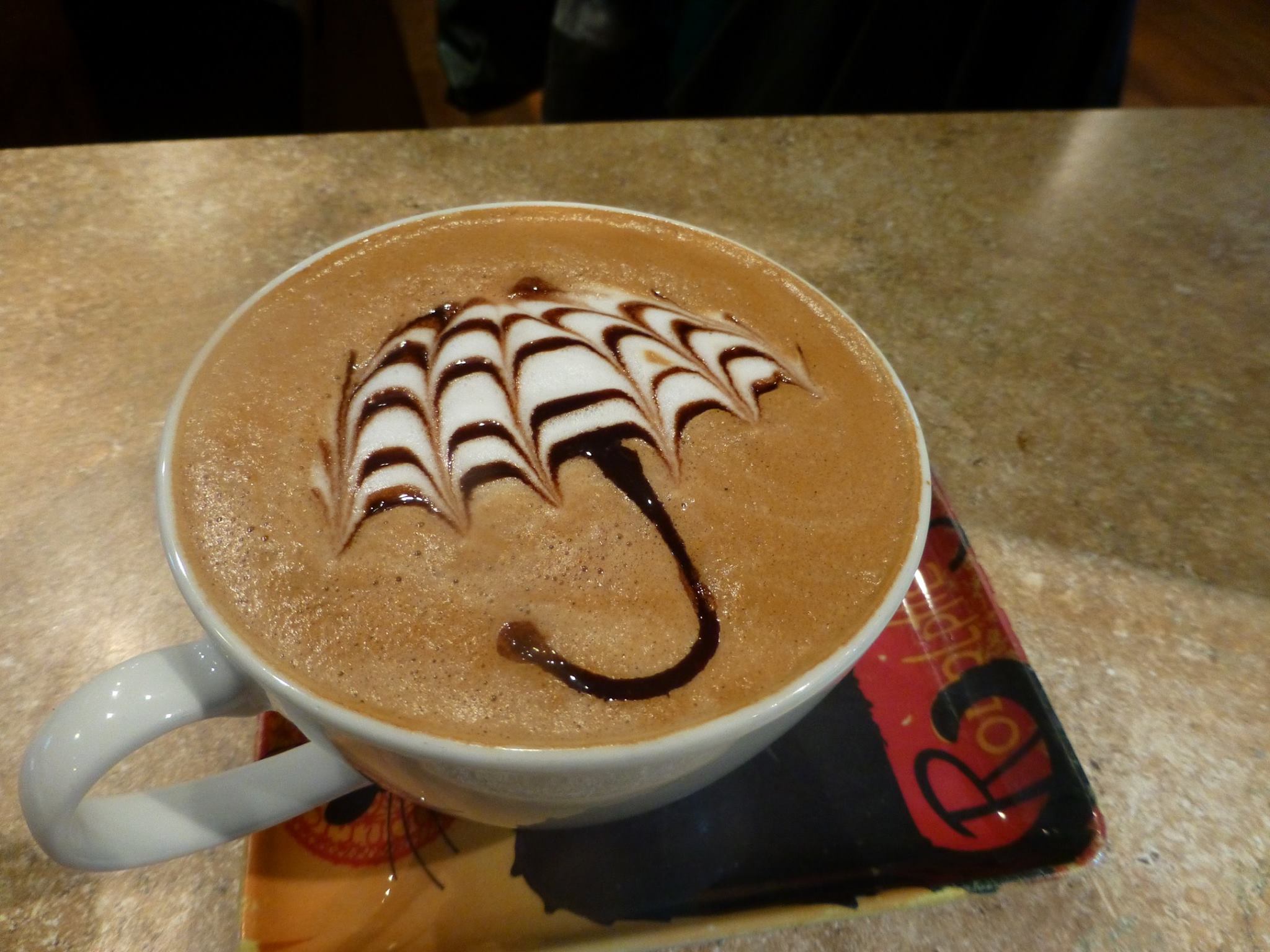 Cup of coffee with whipped cream and chocolate syrup decoration on top, placed on a table with a napkin underneath.