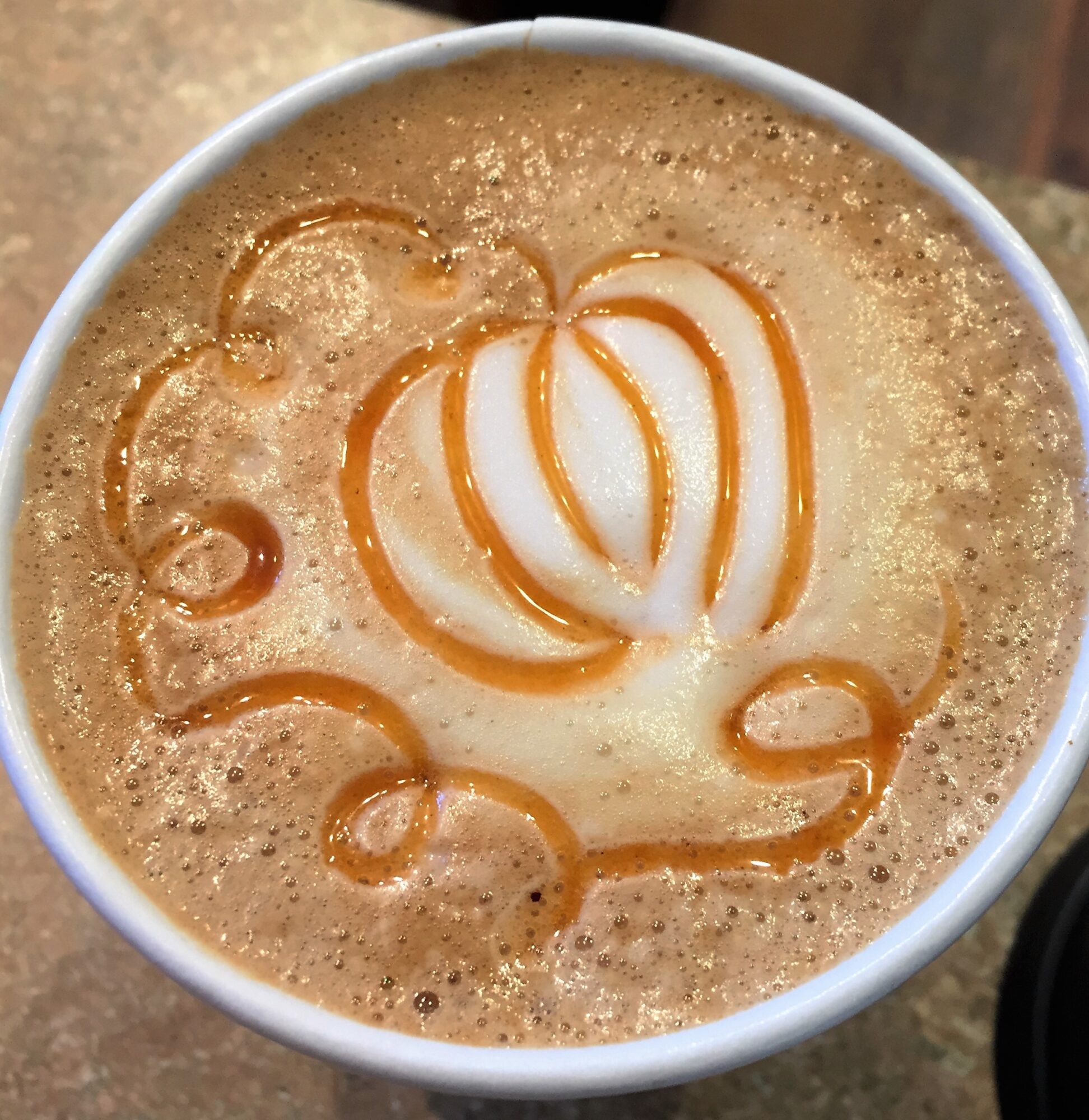 Cup of coffee with foam art resembling a flower, topped with caramel drizzle, viewed from above.