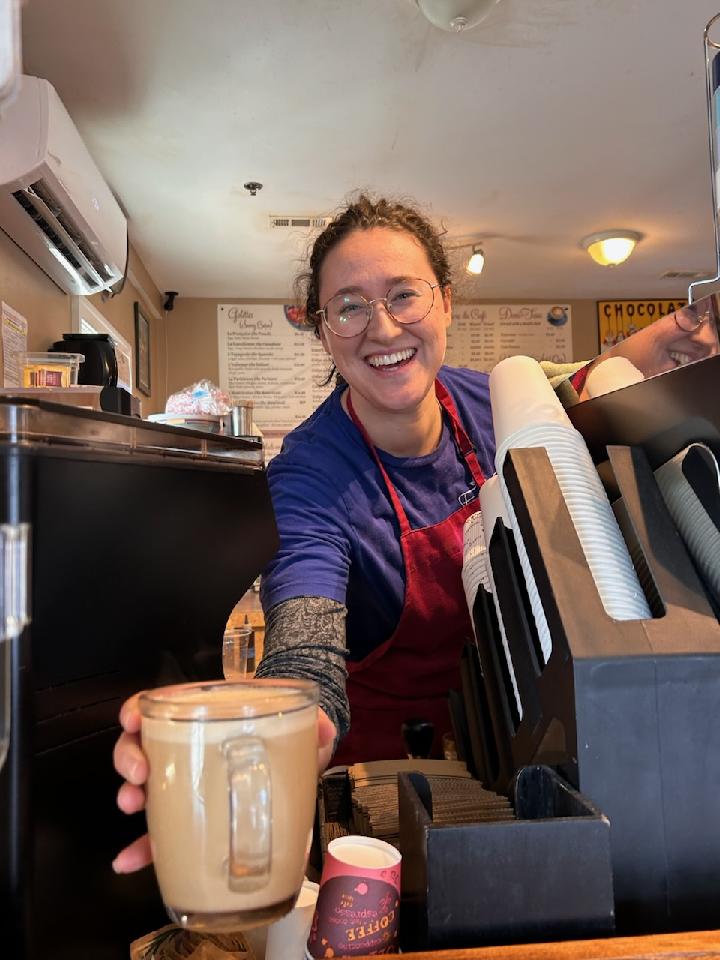 Smiling woman behind a coffee counter, holding a glass of coffee, wearing glasses and a red apron.