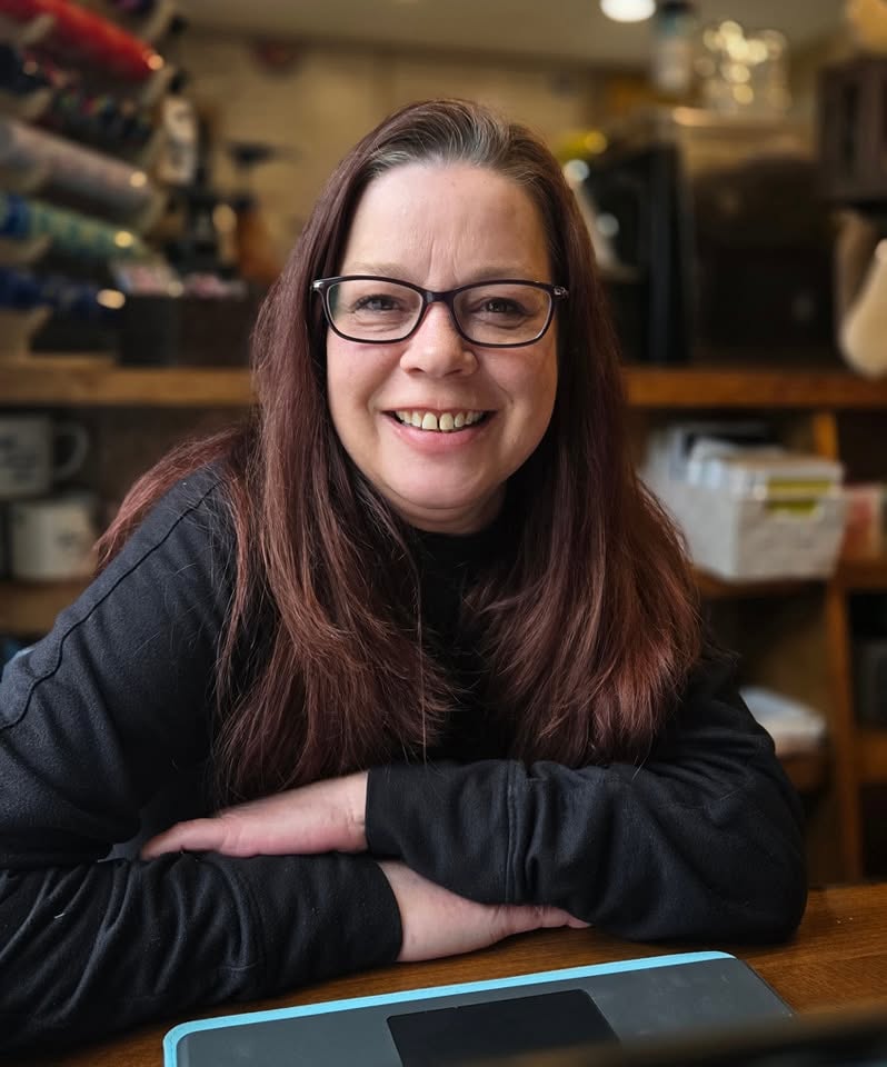 Woman with glasses and long brown hair smiling, sitting at a table in a room with shelves and various items behind her.
