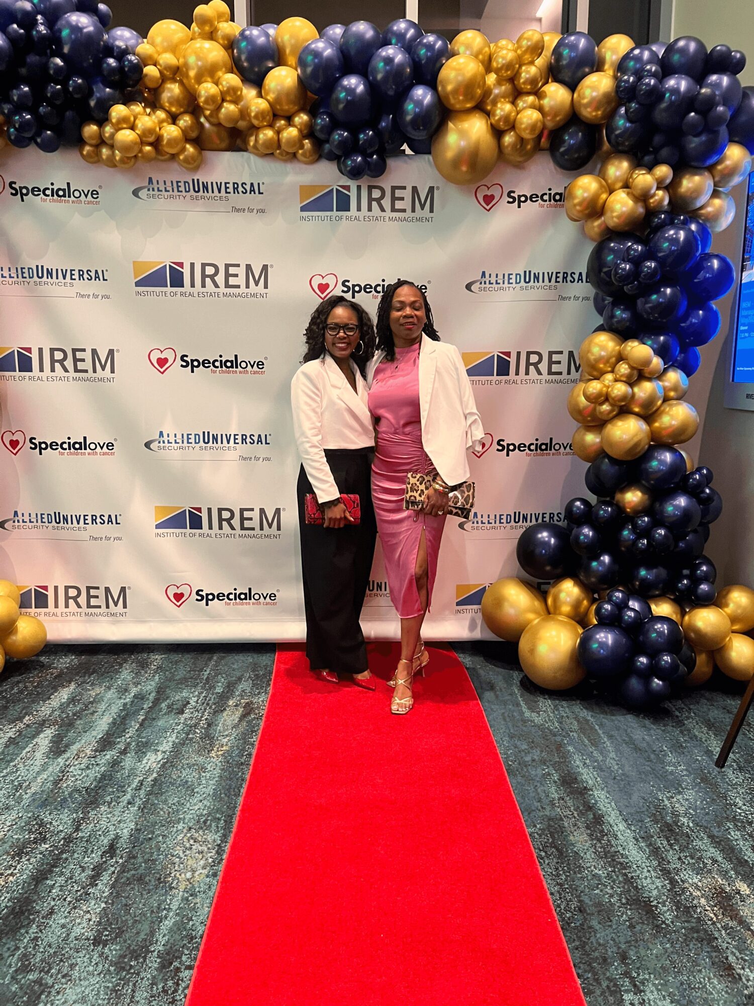 Two women in pink and white outfits stand on a red carpet in front of a decorated backdrop with balloons and logos.