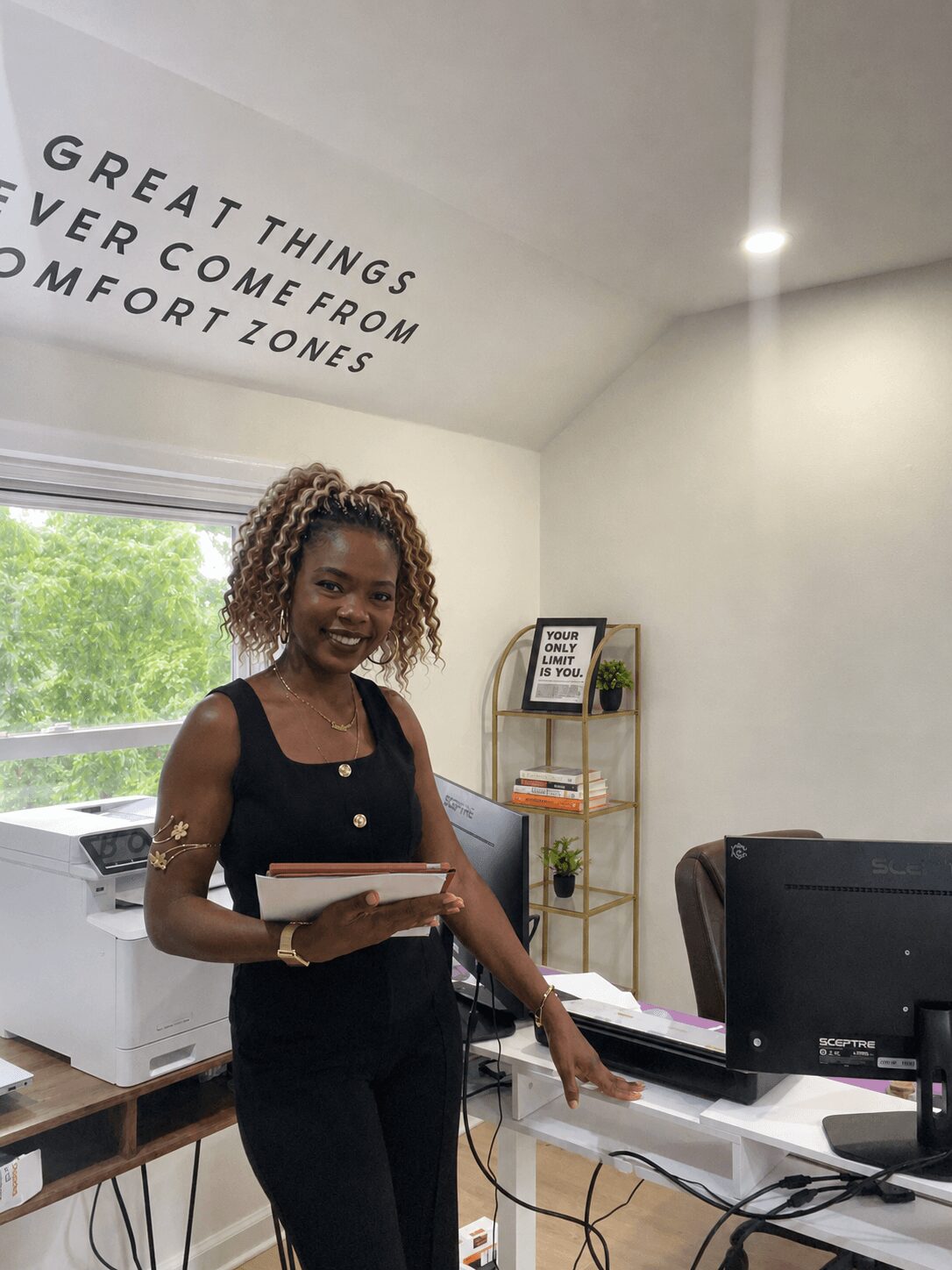 Woman with curly hair holding a tablet in an office, smiling, with a motivational quote on the wall, and a window showing greenery.
