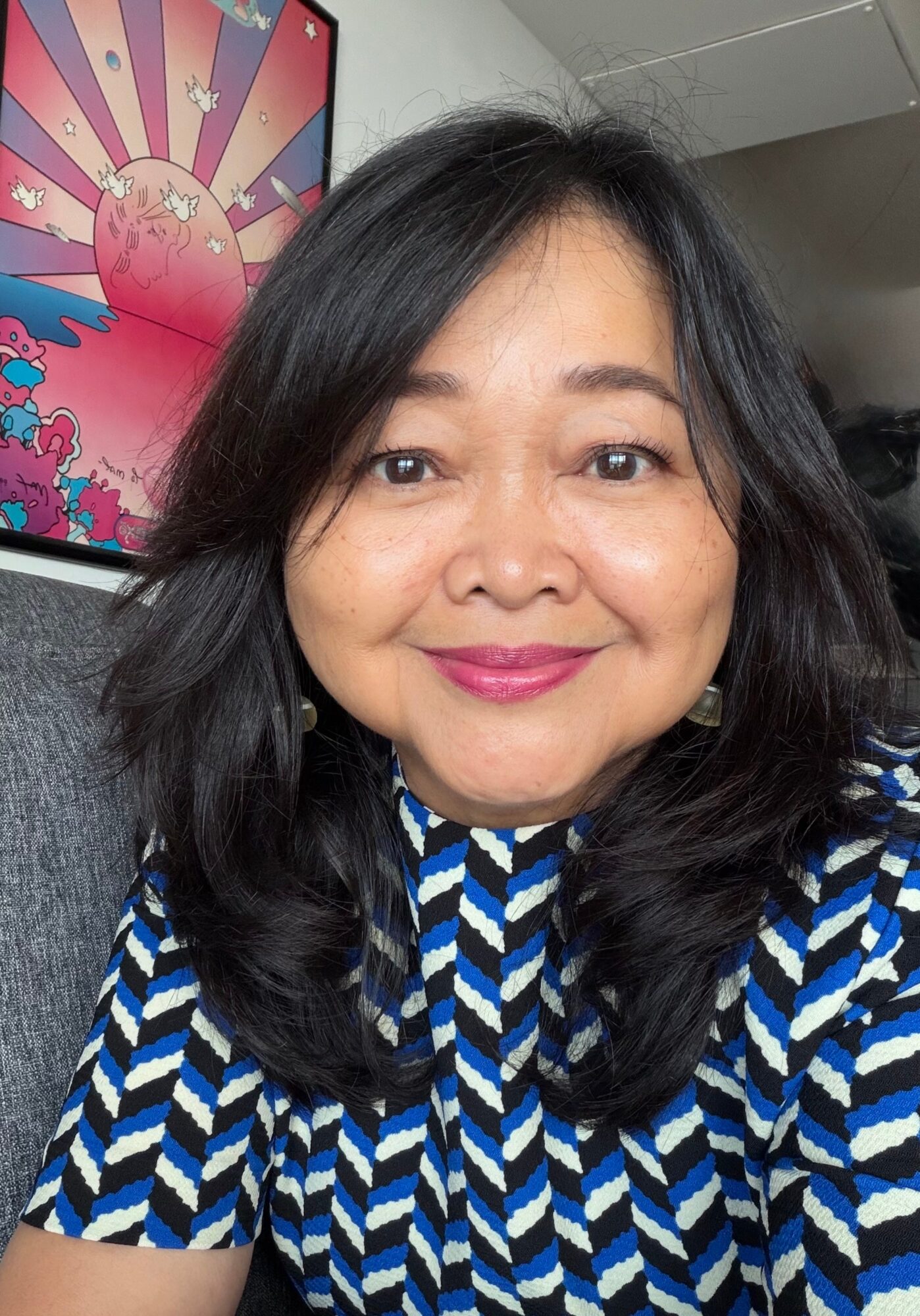 Woman with shoulder-length black hair smiling, wearing a blue and white patterned top, in front of colorful artwork.
