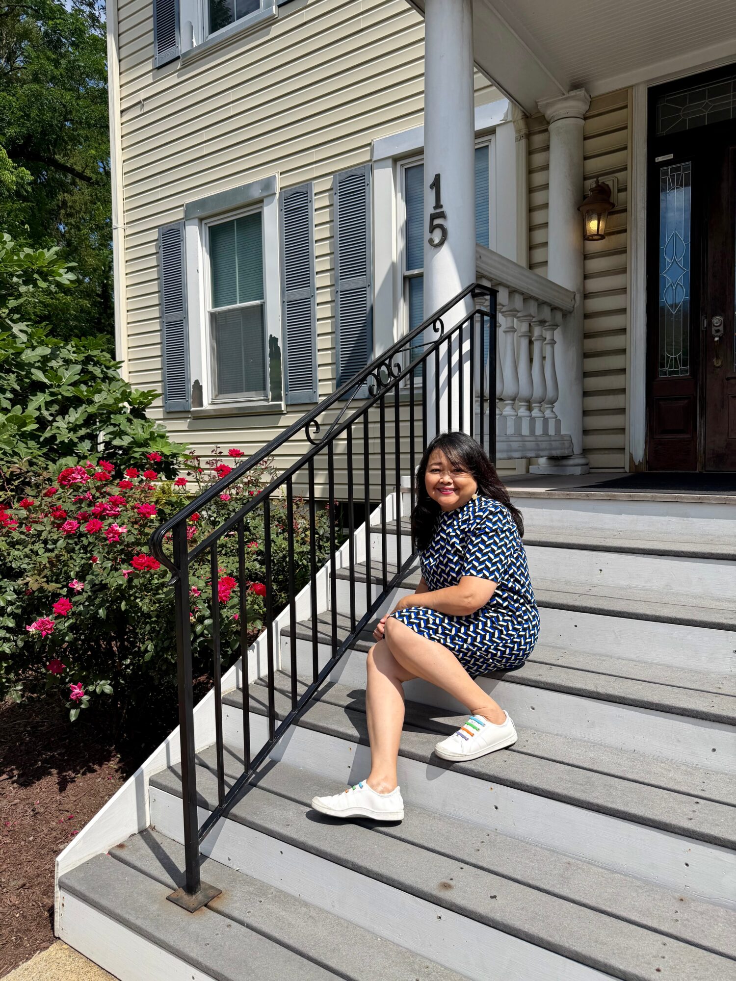 Young woman sitting on front steps of a house, smiling, with flowers and house exterior in background.