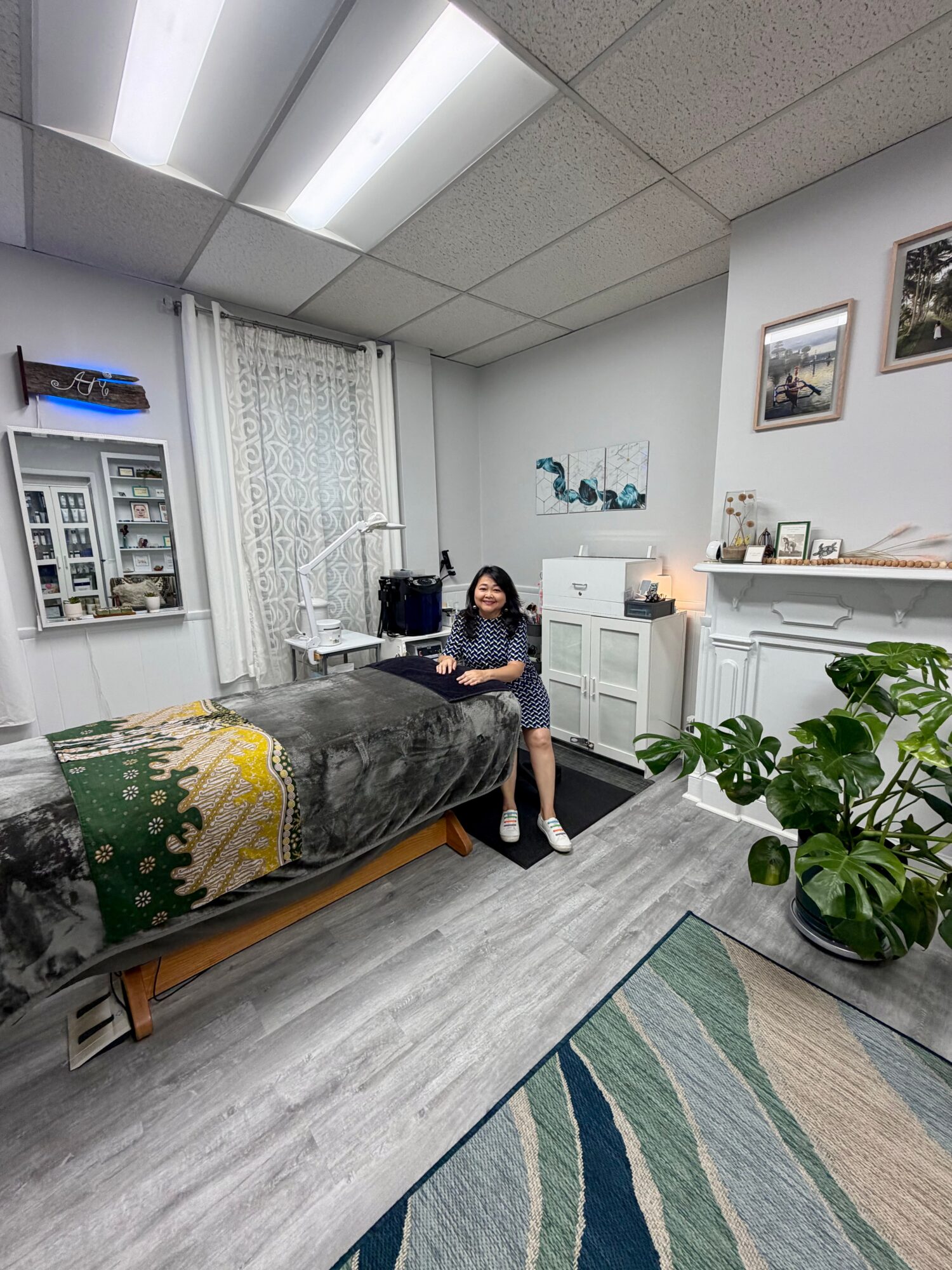 A woman sitting on a bed in a room with white walls, framed pictures, a large plant, and a window with curtains.