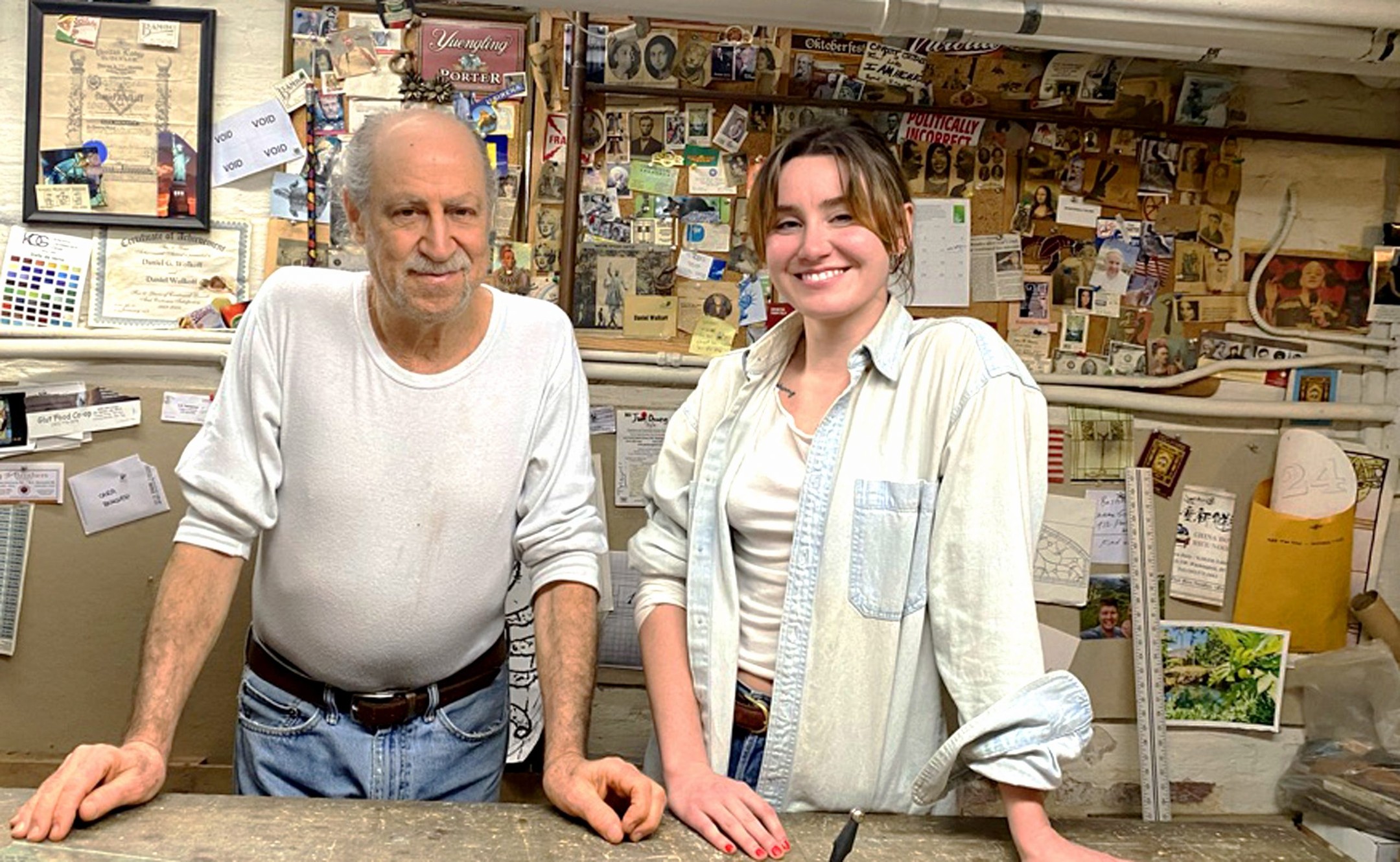 Two people standing behind a counter in a store, smiling, with shelves and items in the background.