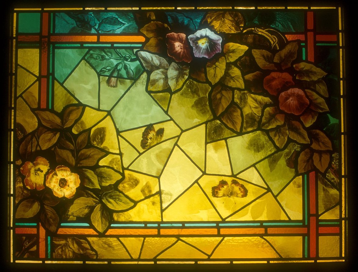 Colorful stained glass window with flowers and leaves, featuring yellow, purple, and green hues, framed by red and green borders.