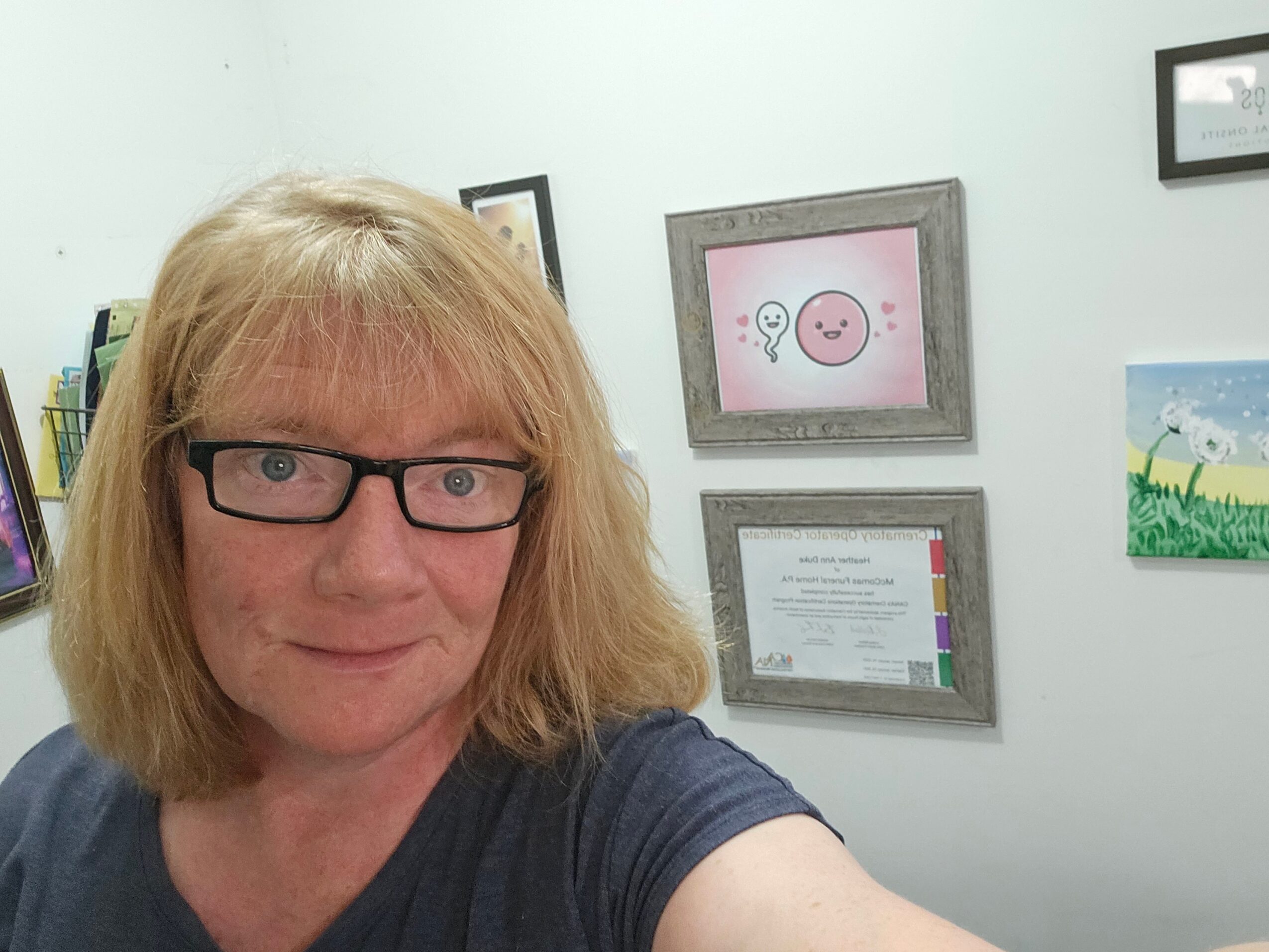 Woman with glasses and blonde hair taking a selfie in a room with framed pictures on the wall.