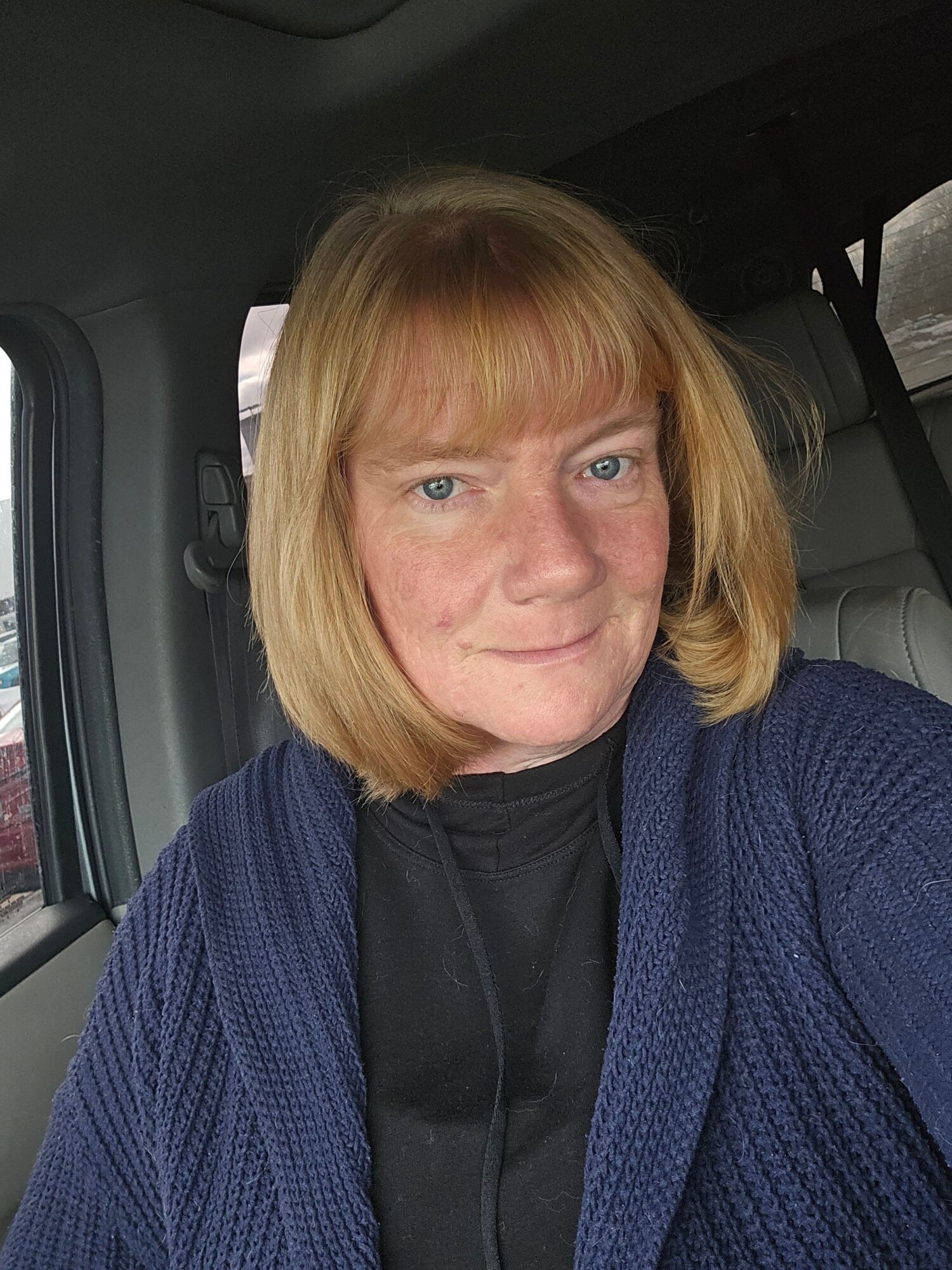 Woman with blonde hair and blue eyes sitting in a vehicle, wearing a dark top and blue cardigan.