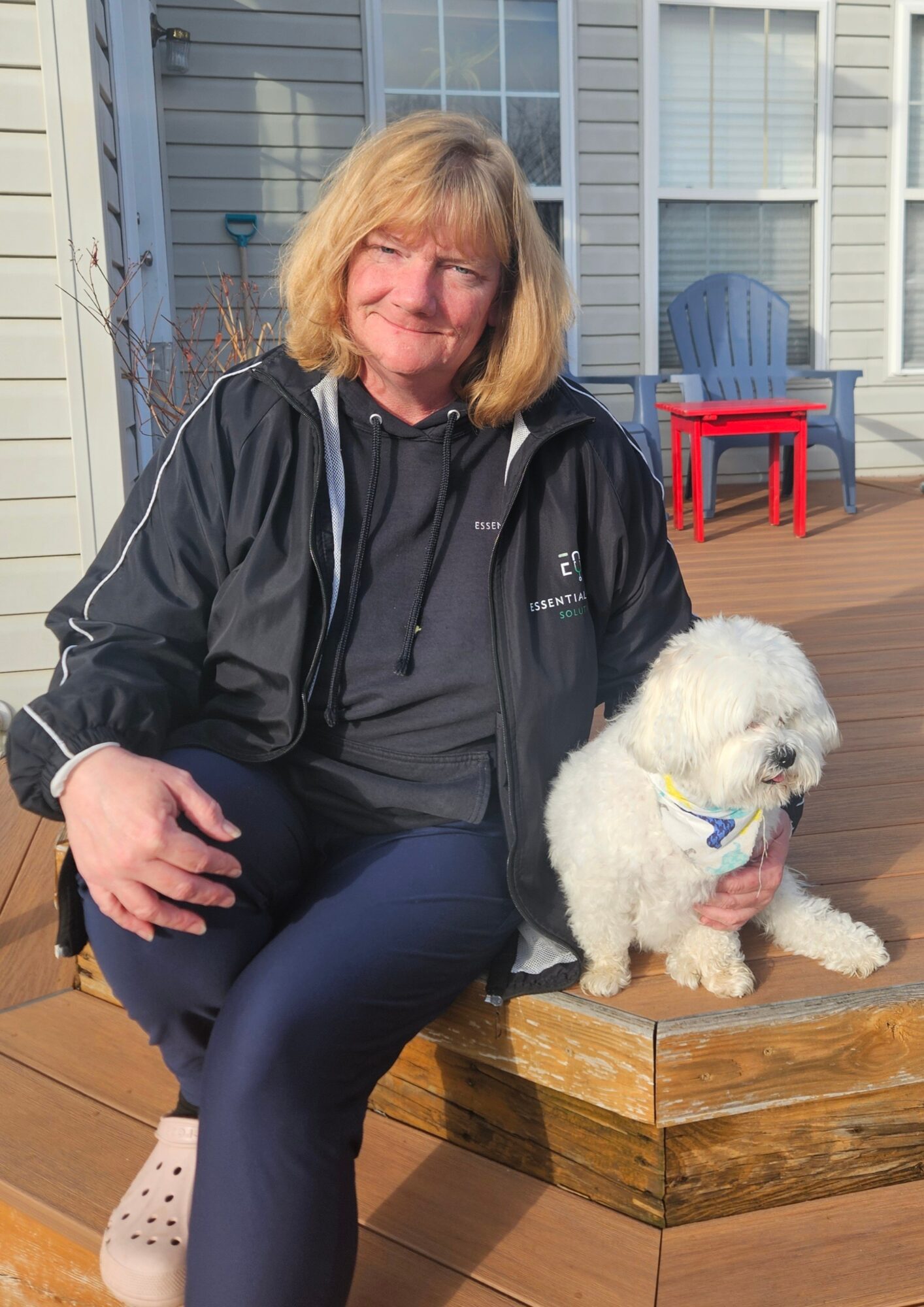 Woman with blonde hair sitting outdoors with a white fluffy dog on a wooden deck, house and chairs in background.