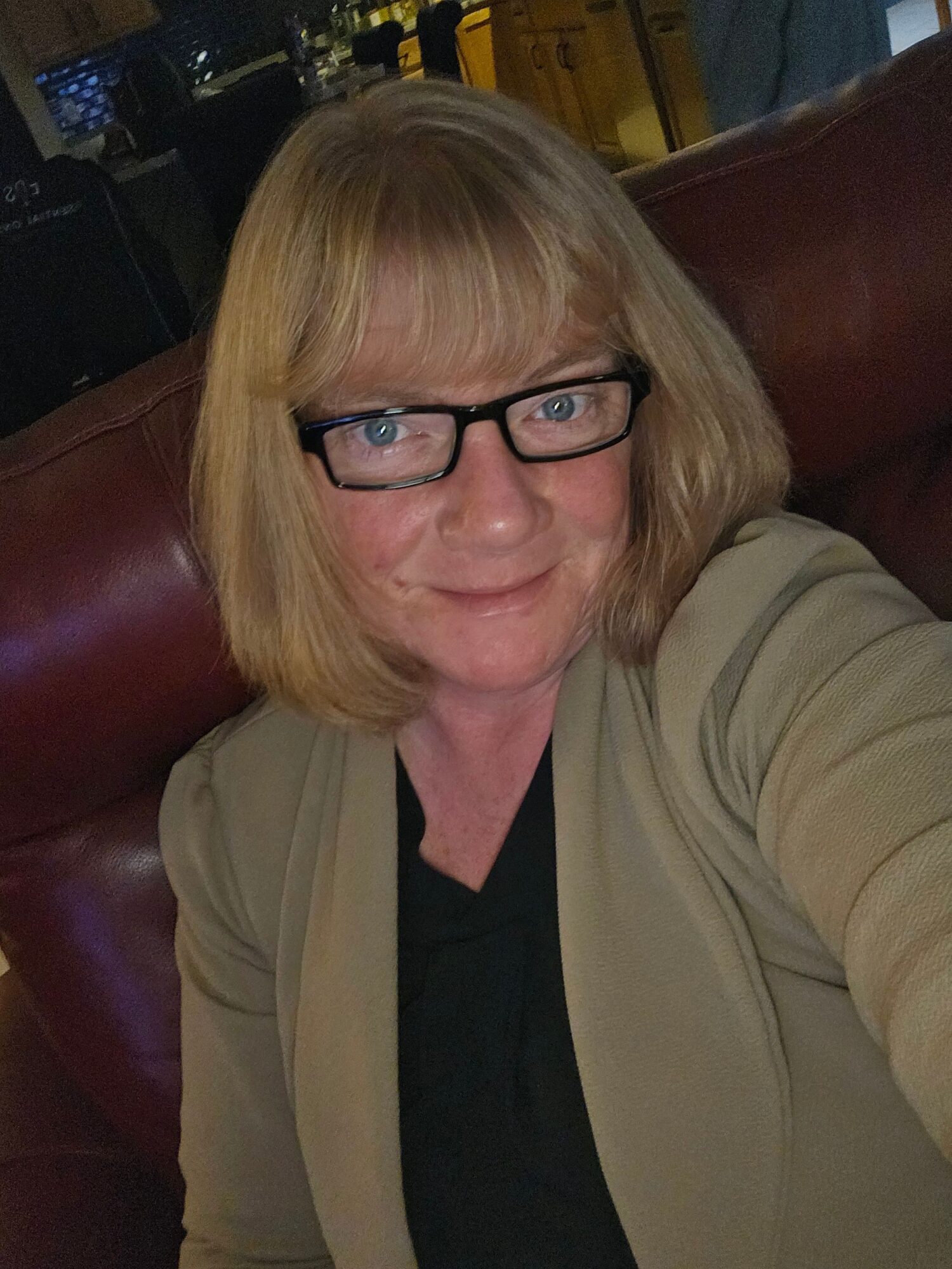 Woman with blonde hair and glasses sitting on a red leather chair, smiling at the camera.