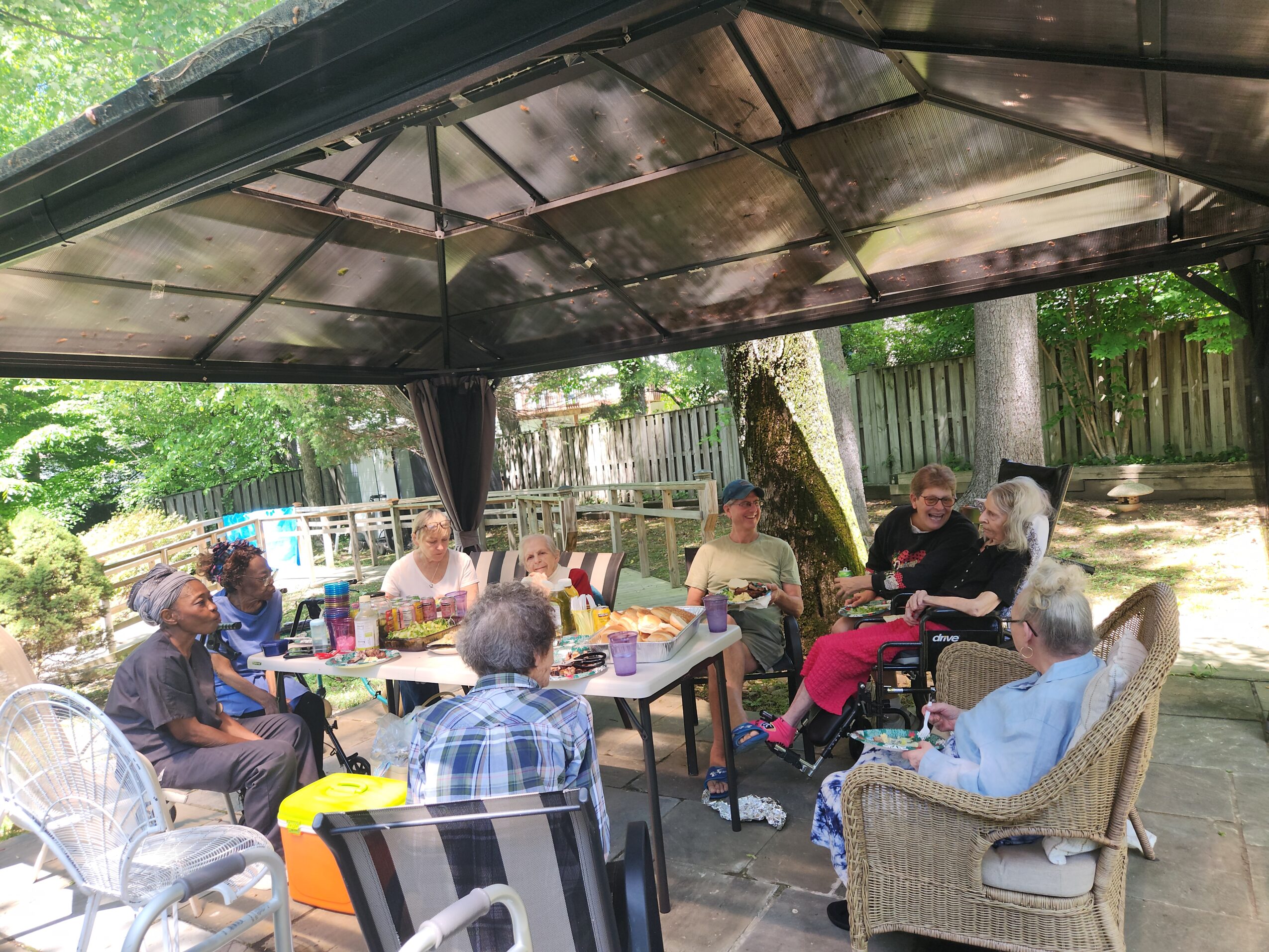 Group of people sitting under a canopy outdoors, socializing around tables with drinks and snacks.