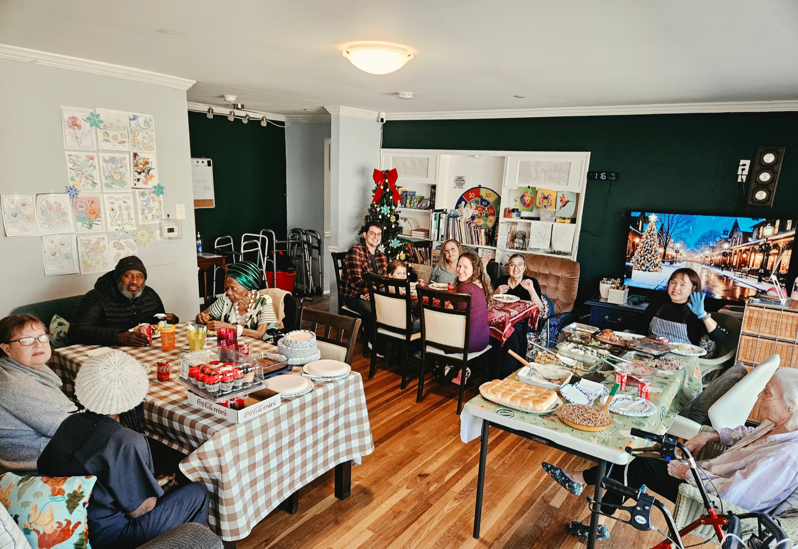 People gathered around tables with food and drinks in a decorated room, some watching a television, festive decorations present.