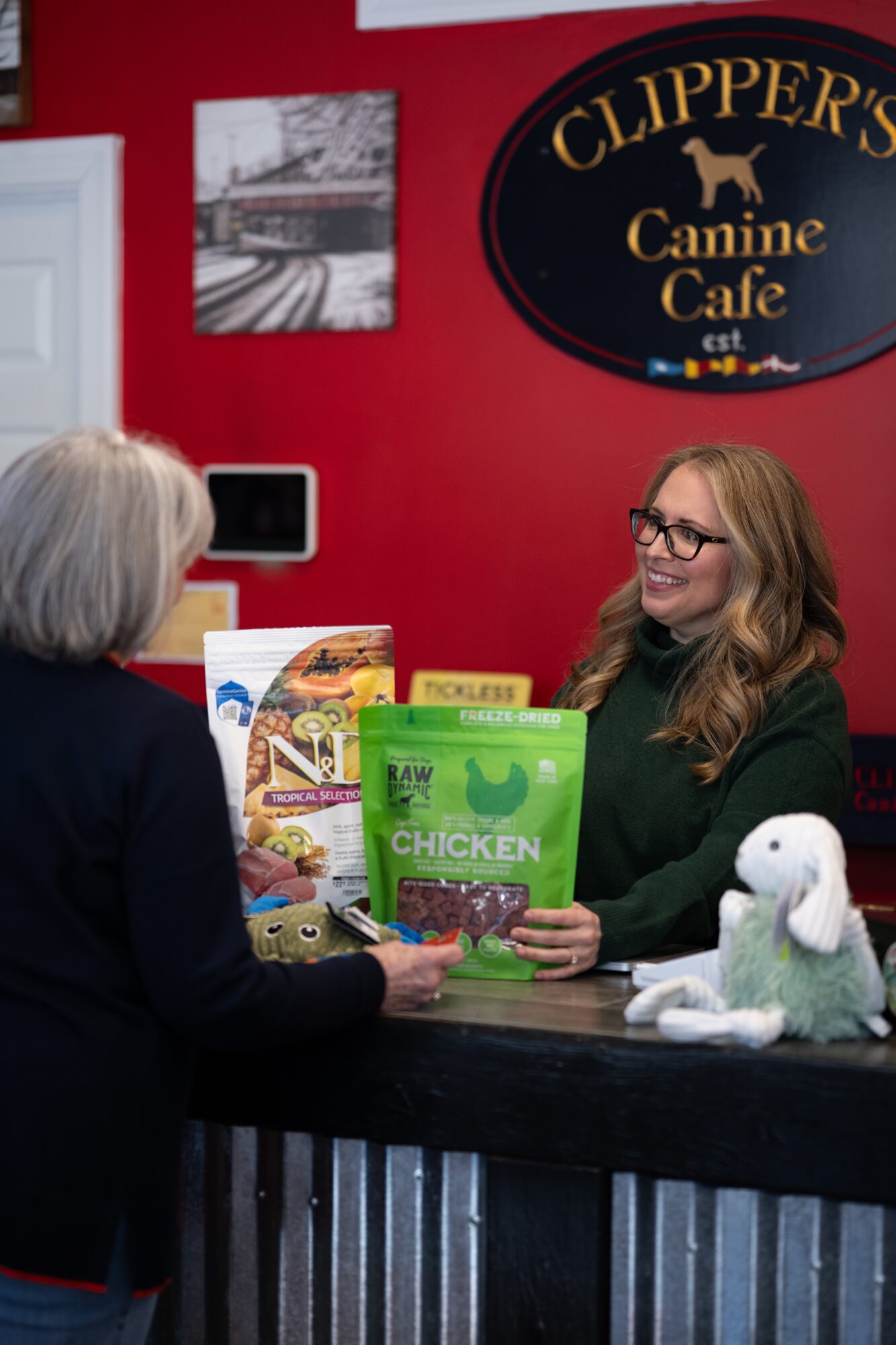 Two women at a counter inside a pet cafe, with a sign reading 'Clippers Canine Cafe' on a red wall, and a plush dog toy on the counter.