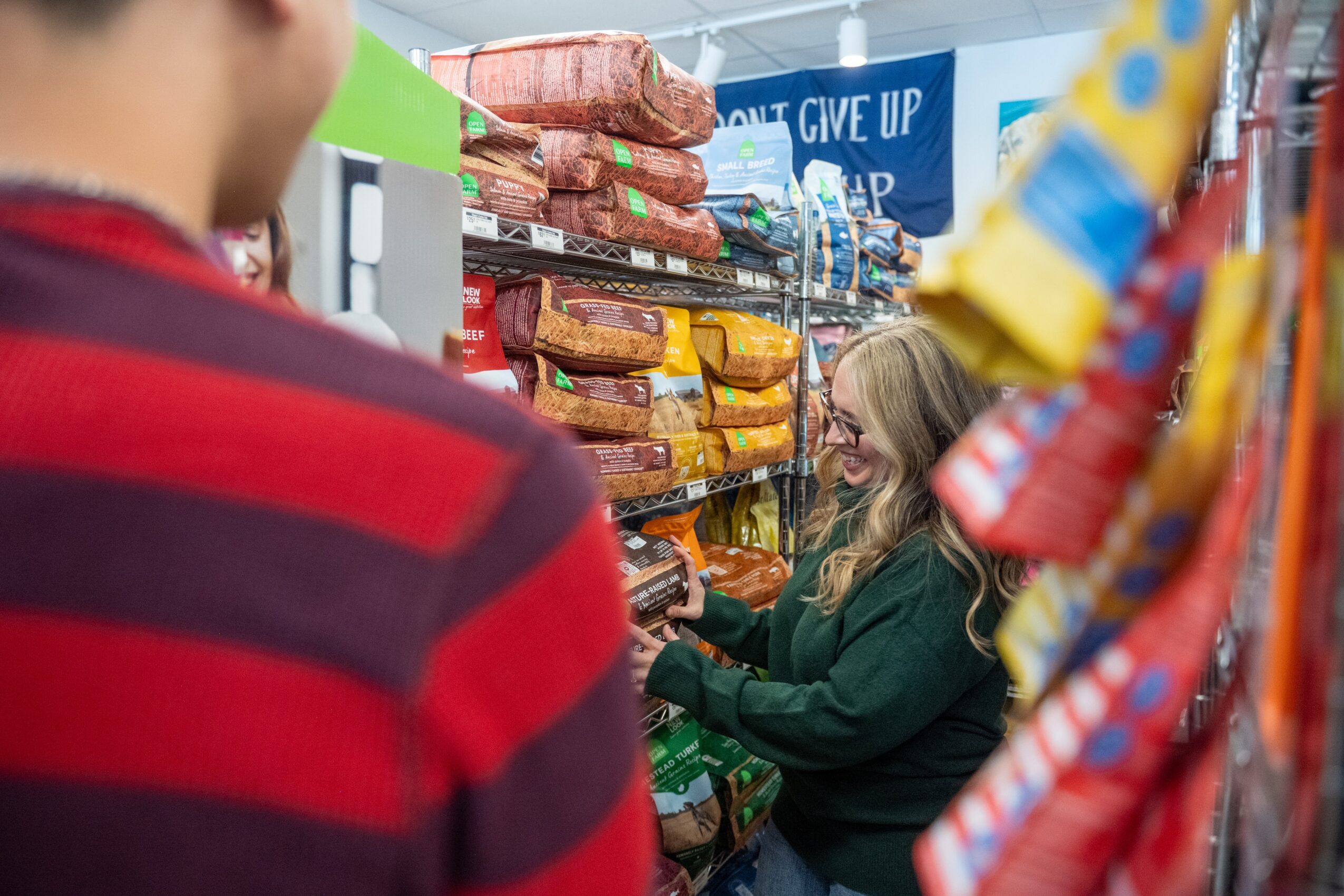 A woman shopping in a store aisle with snacks and colorful items, seen from behind a person in a red and black striped shirt.
