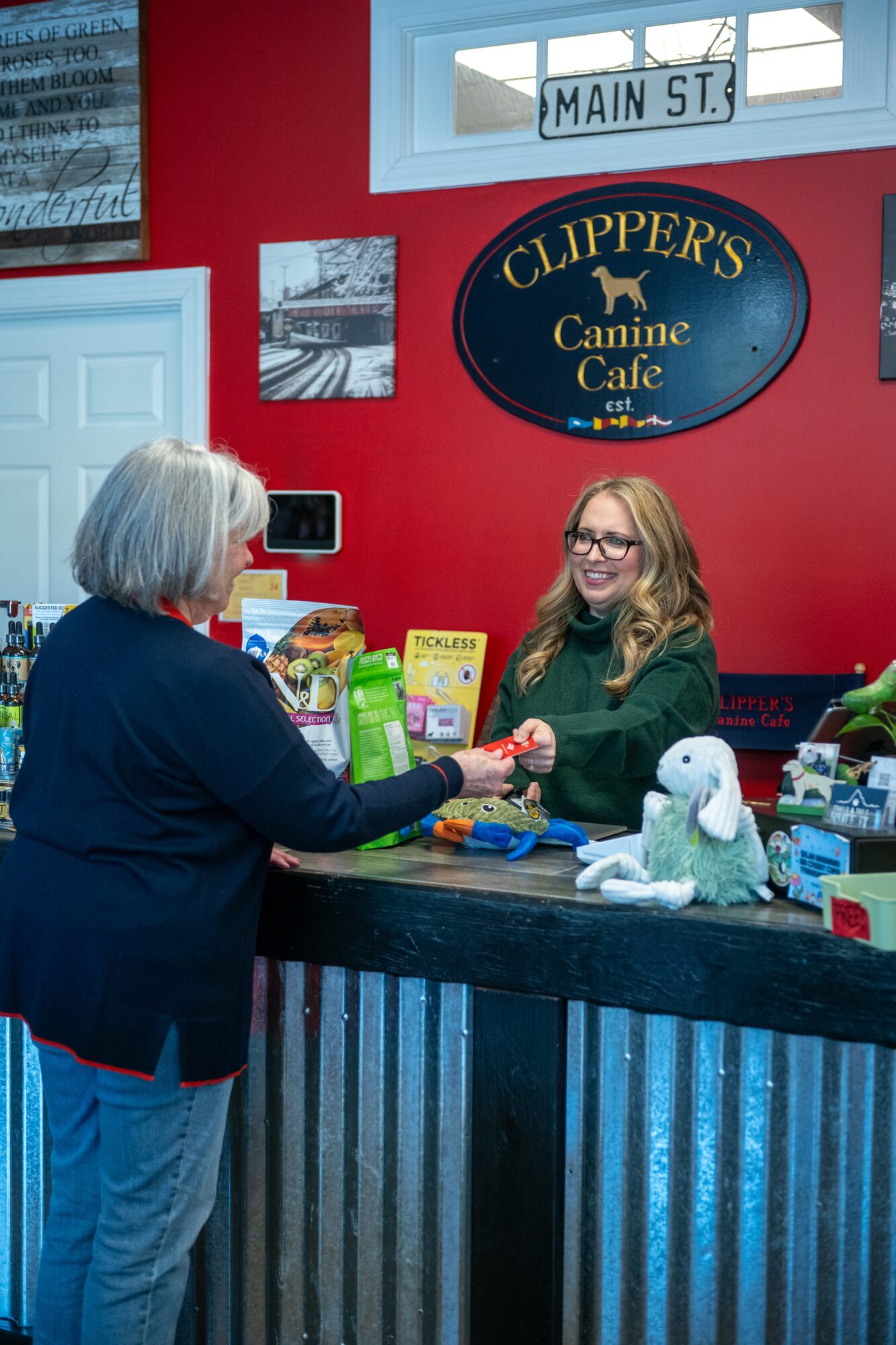 Woman and girl shake hands at a counter inside a pet cafe with a sign reading 'Clippers Canine Cafe' on a red wall.