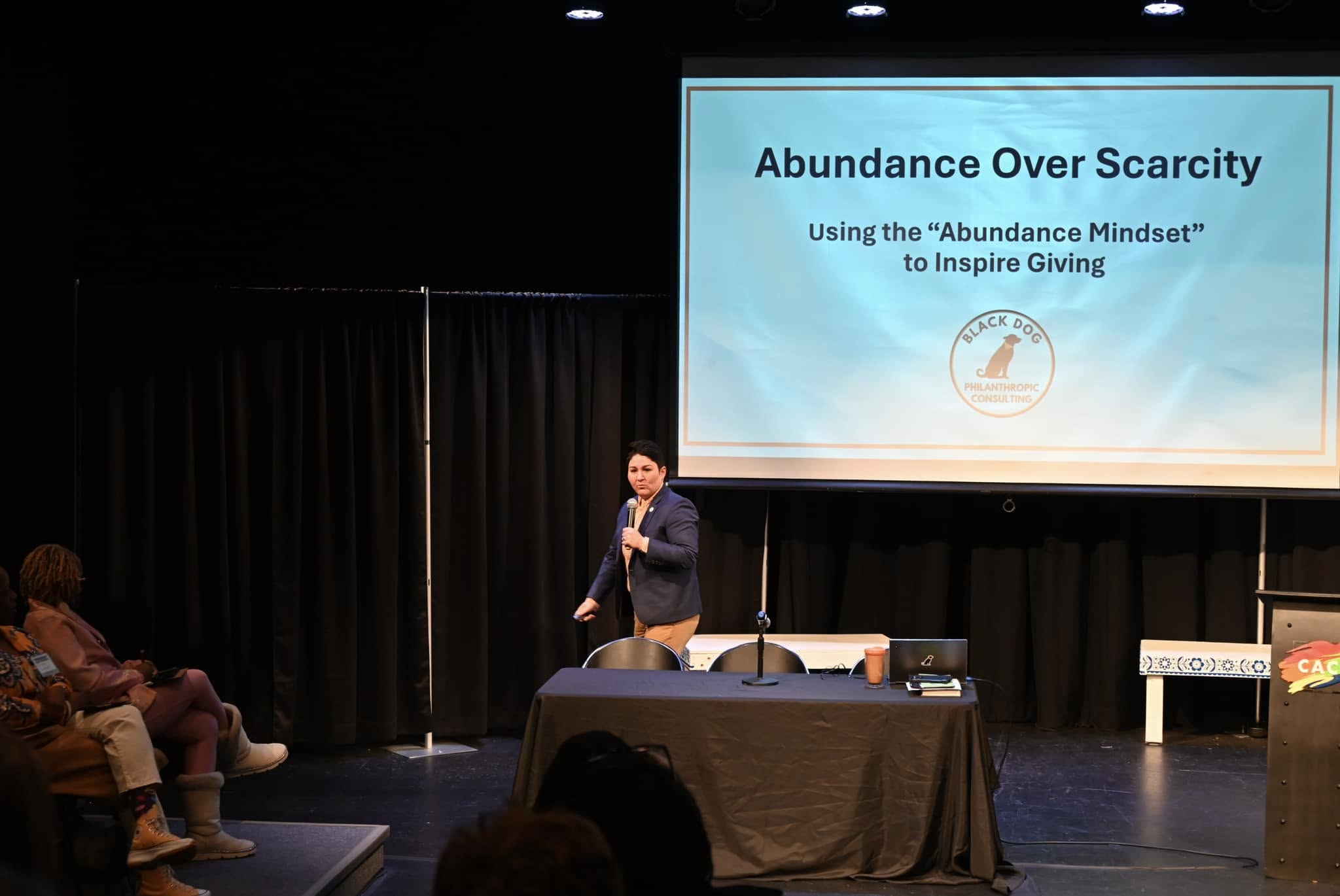 Speaker presenting on stage with a large screen displaying a presentation titled 'Abundance Over Scarcity' in front of an audience.