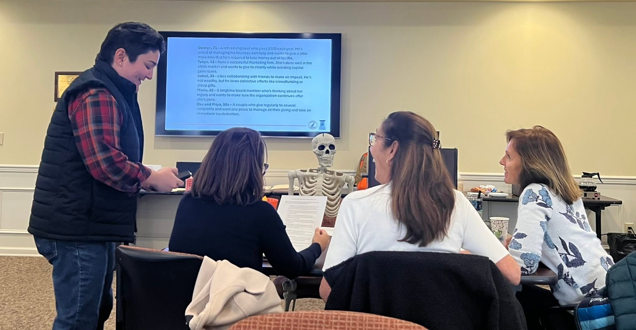Person standing and speaking to four seated people in a classroom with a large screen and a skeleton model.