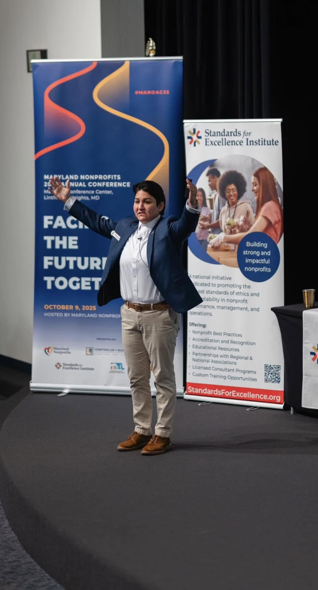 Man in a suit standing on stage with arms raised, two banners behind him, one with blue and red design, another with group photo.