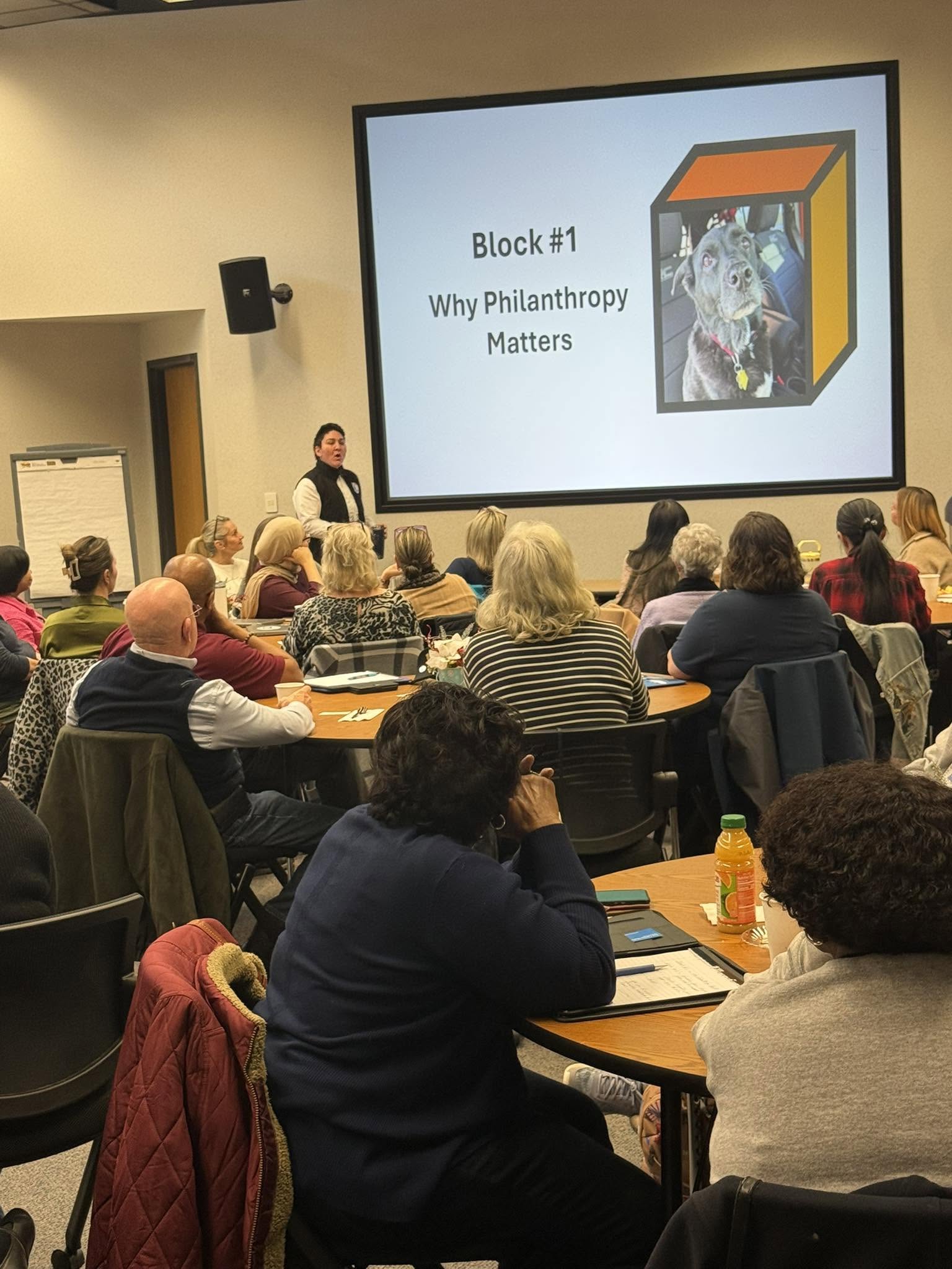 A woman presents in front of a large screen displaying a slide titled 'Block #1 Why Philanthropy Matters' at a conference room with seated attendees.