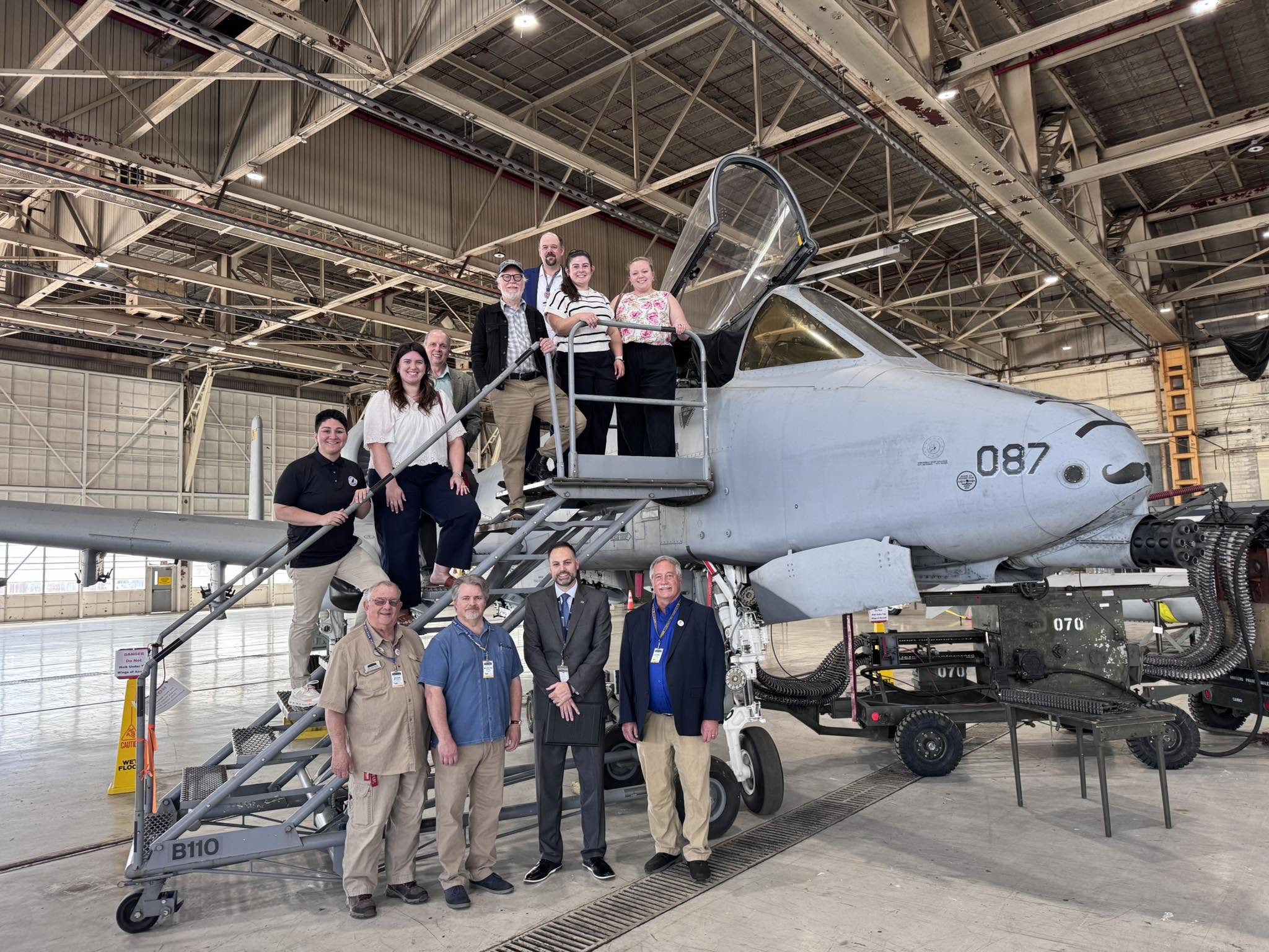 Group of people standing in front of a military aircraft inside a hangar, with some on stairs and others on the ground.