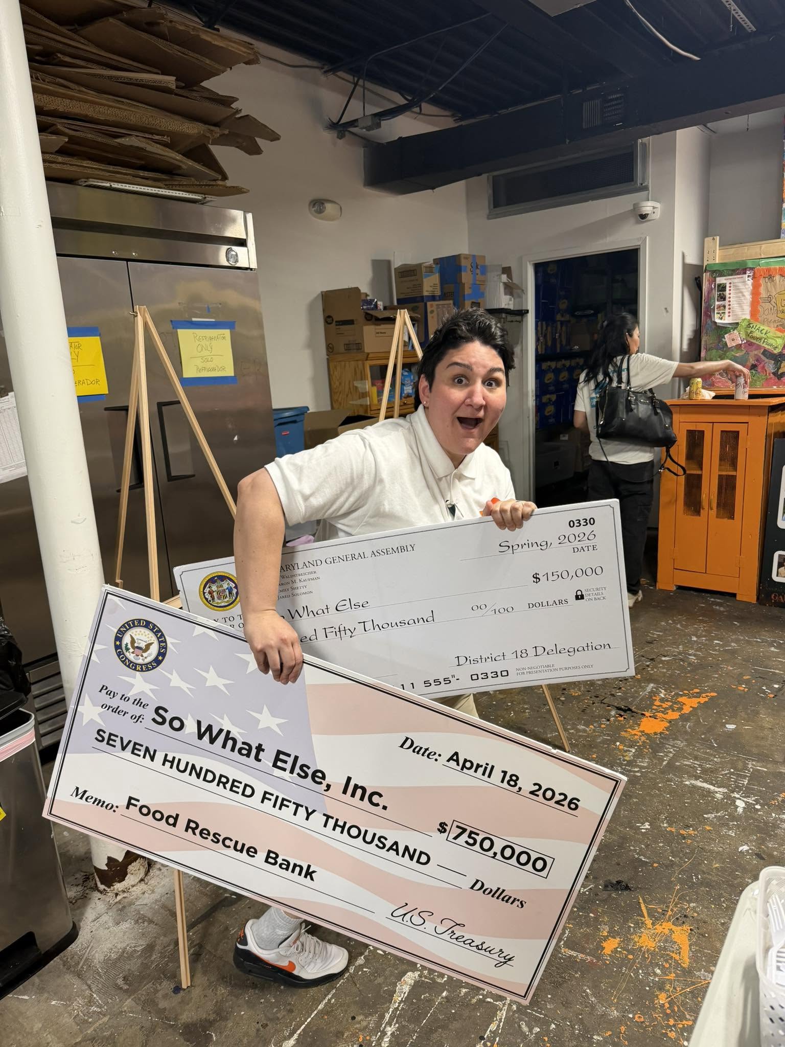 Woman holding a large check in a warehouse or storage area, smiling and looking at the camera.