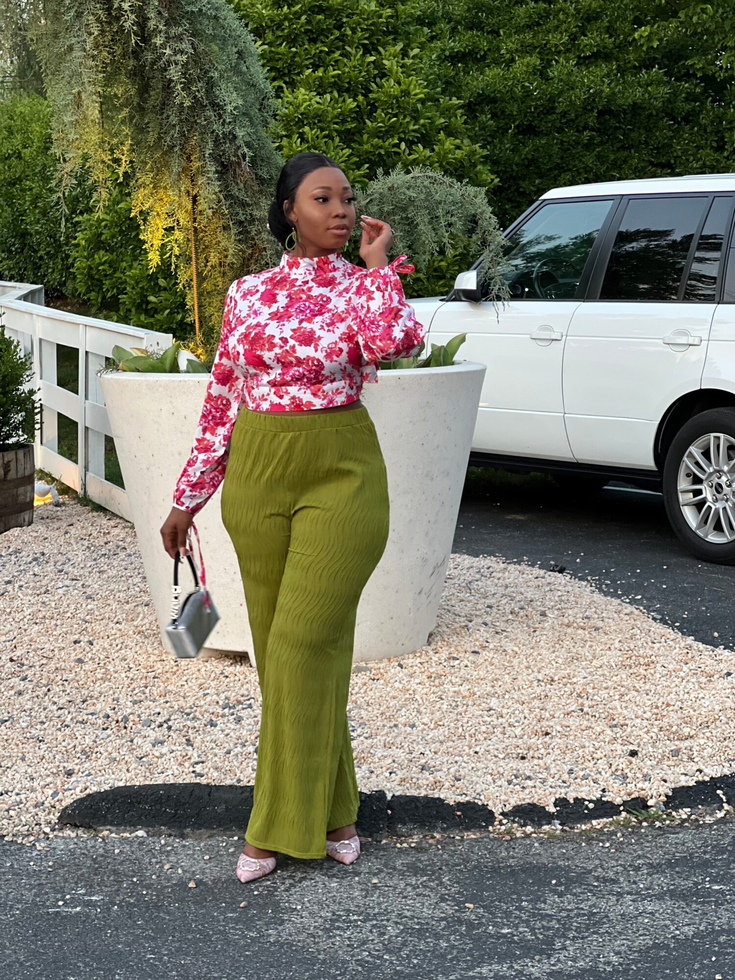 Woman in floral blouse and green pants standing outdoors near a white vehicle and large planter, with trees in background.