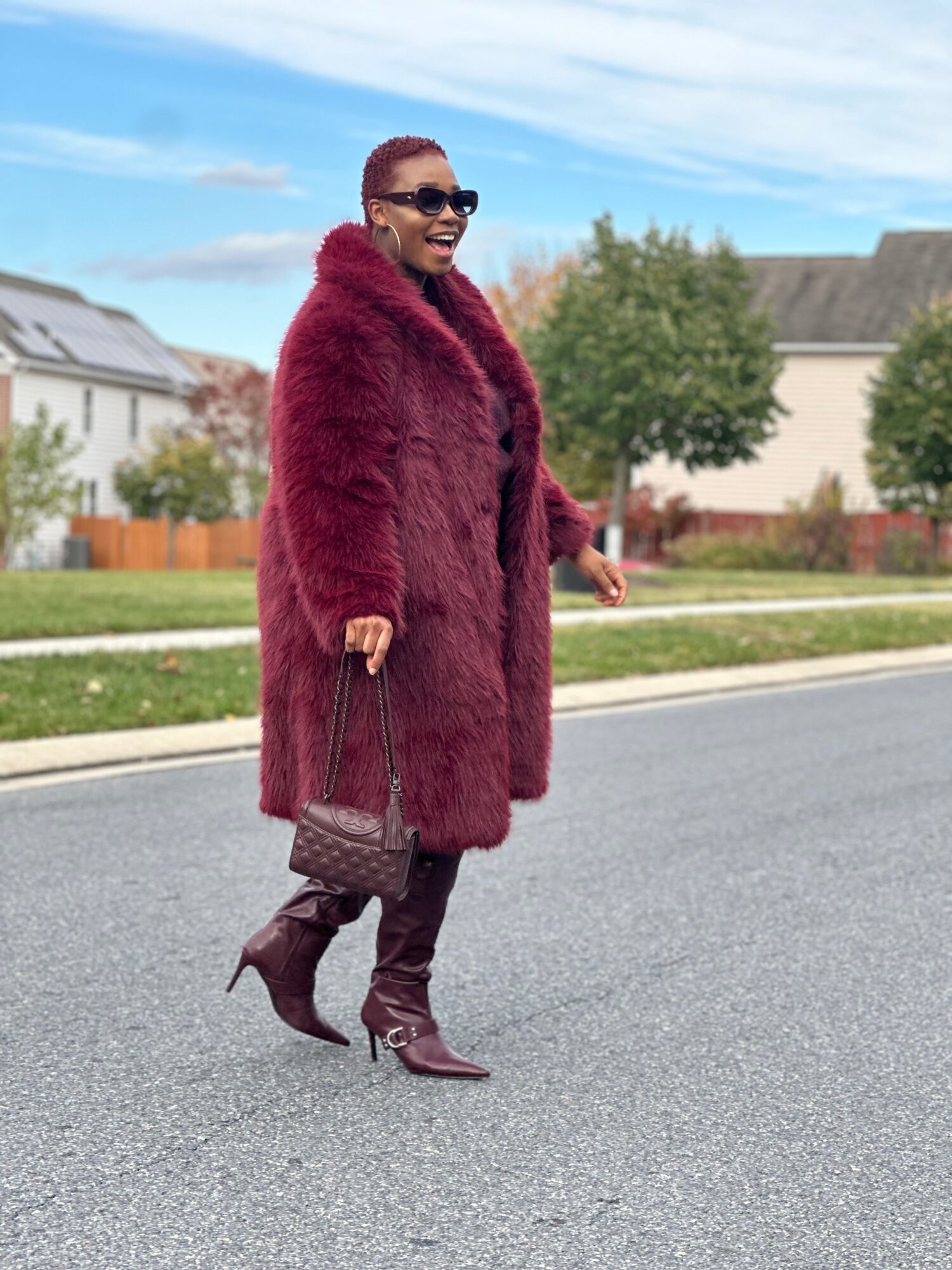 Woman walking on street wearing a red fur coat, sunglasses, and high-heeled boots, holding a purse, smiling, with houses and trees in background.