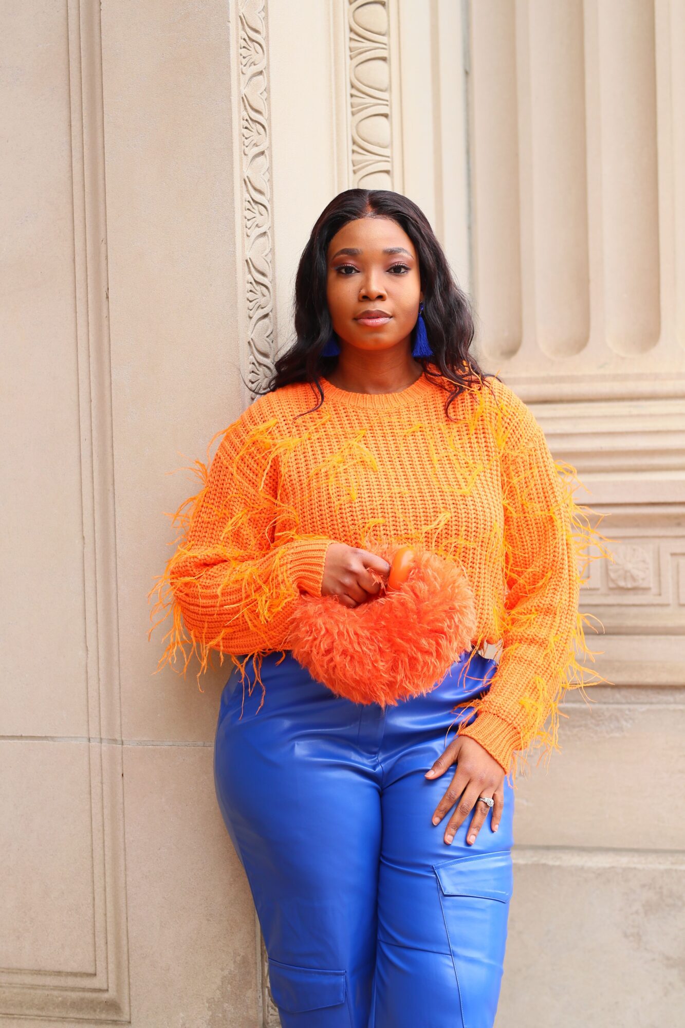 Woman in orange sweater and blue pants standing indoors with decorative wall details.