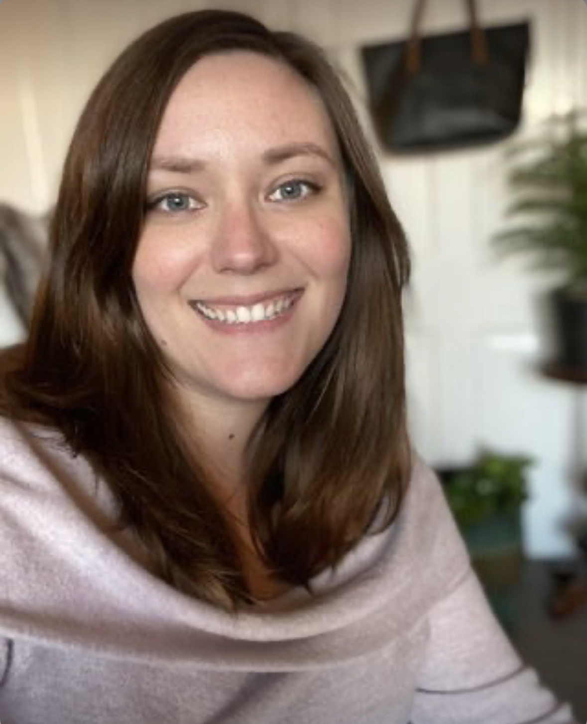 Young woman with brown hair smiling indoors, background includes a TV and plants.
