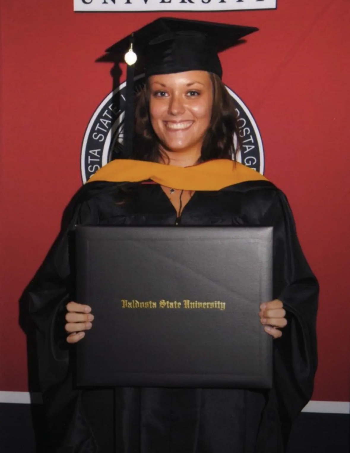 Young woman in graduation cap and gown holding diploma case, smiling, red background with university emblem, text on diploma case, 20 words.