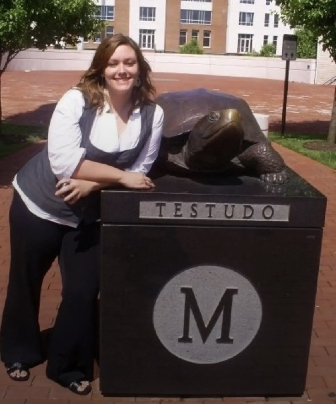 Woman smiling next to a turtle statue on a pedestal with 'TESTUDO' and 'M' symbols, outdoors on a sunny day.