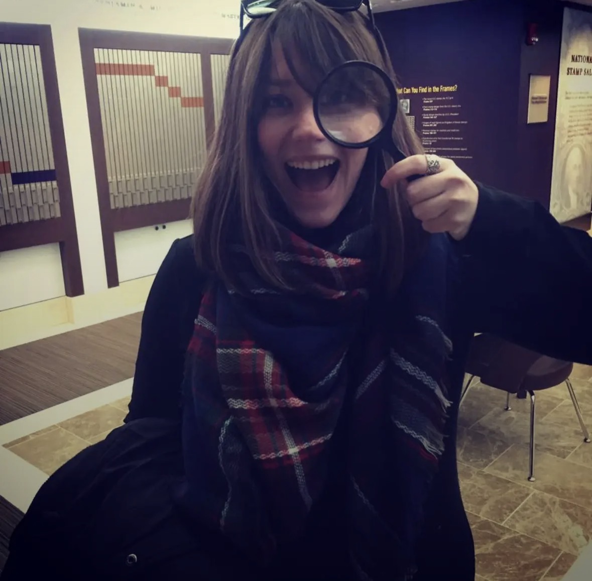 Person smiling and holding a magnifying glass in front of one eye, indoors with chairs and informational displays.