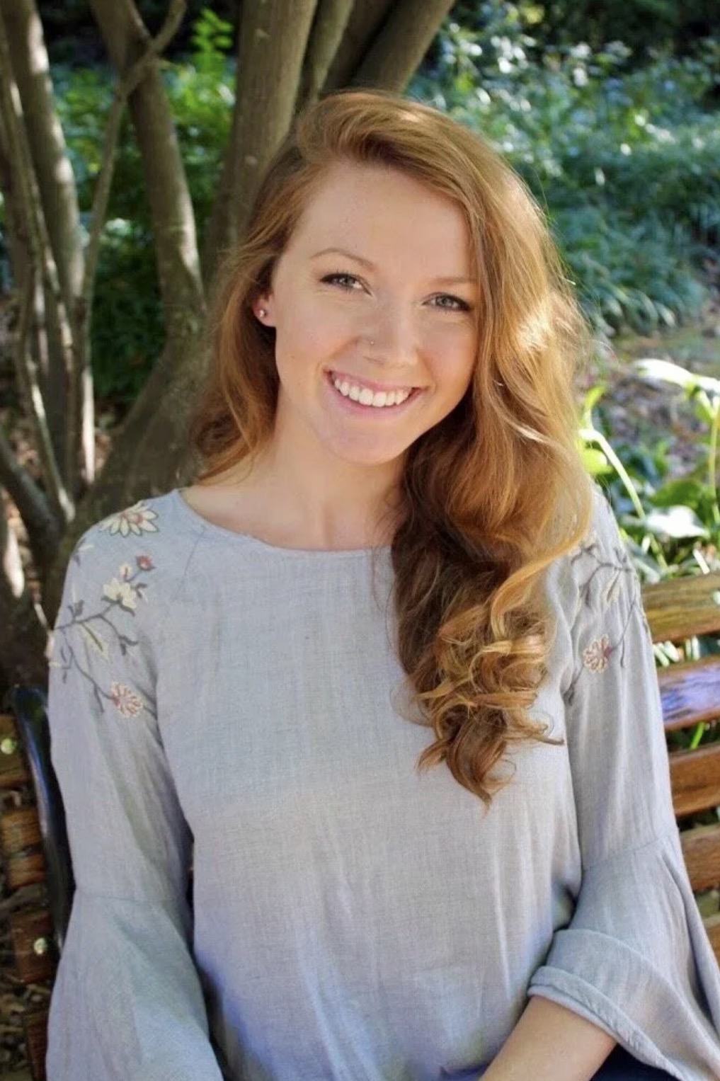 Young woman with long, wavy red hair smiling outdoors, wearing a light gray blouse with embroidered details, surrounded by greenery.