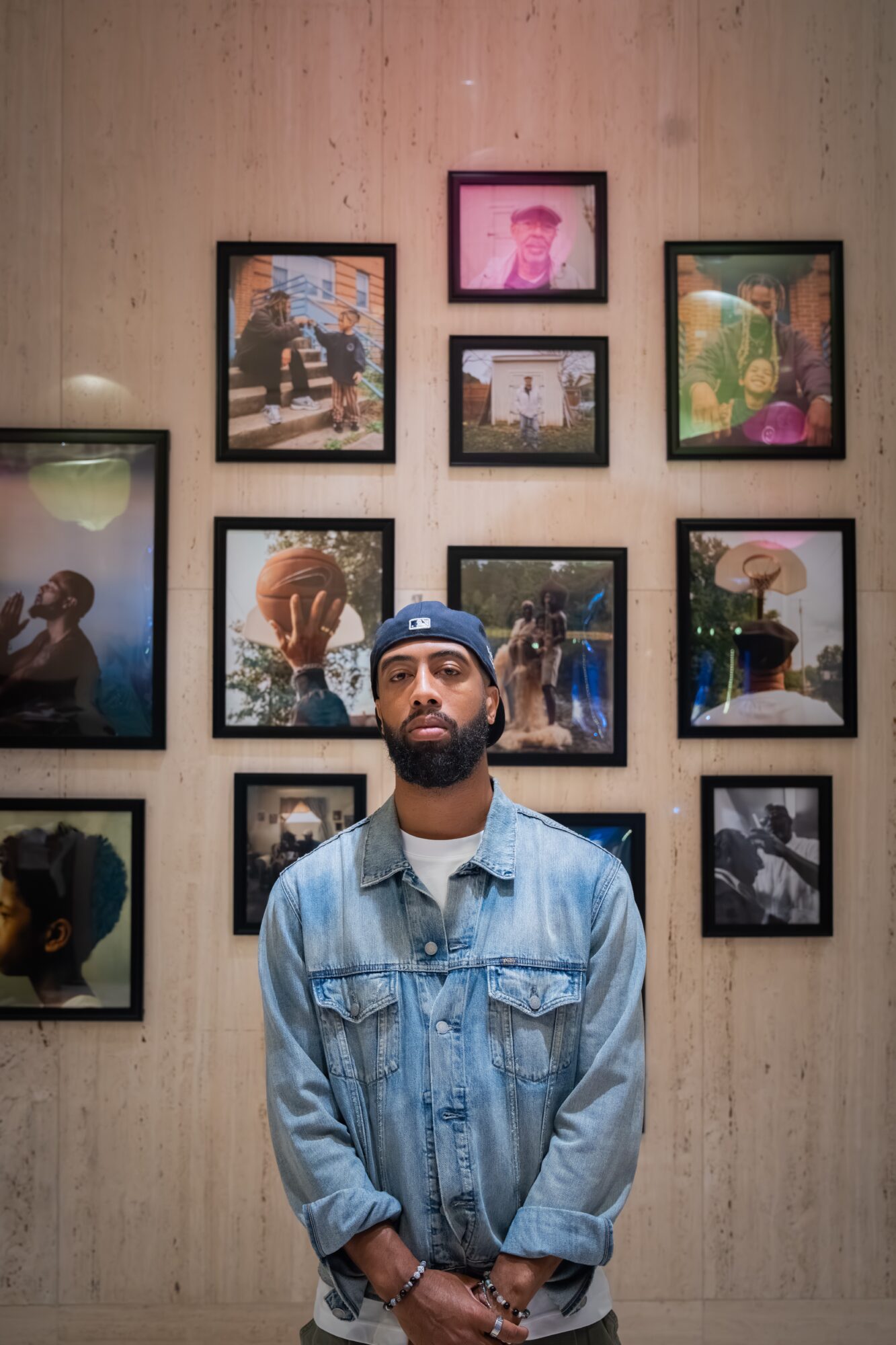 Man with a beard and denim jacket standing in front of a wall with framed photographs, some with people and outdoor scenes.