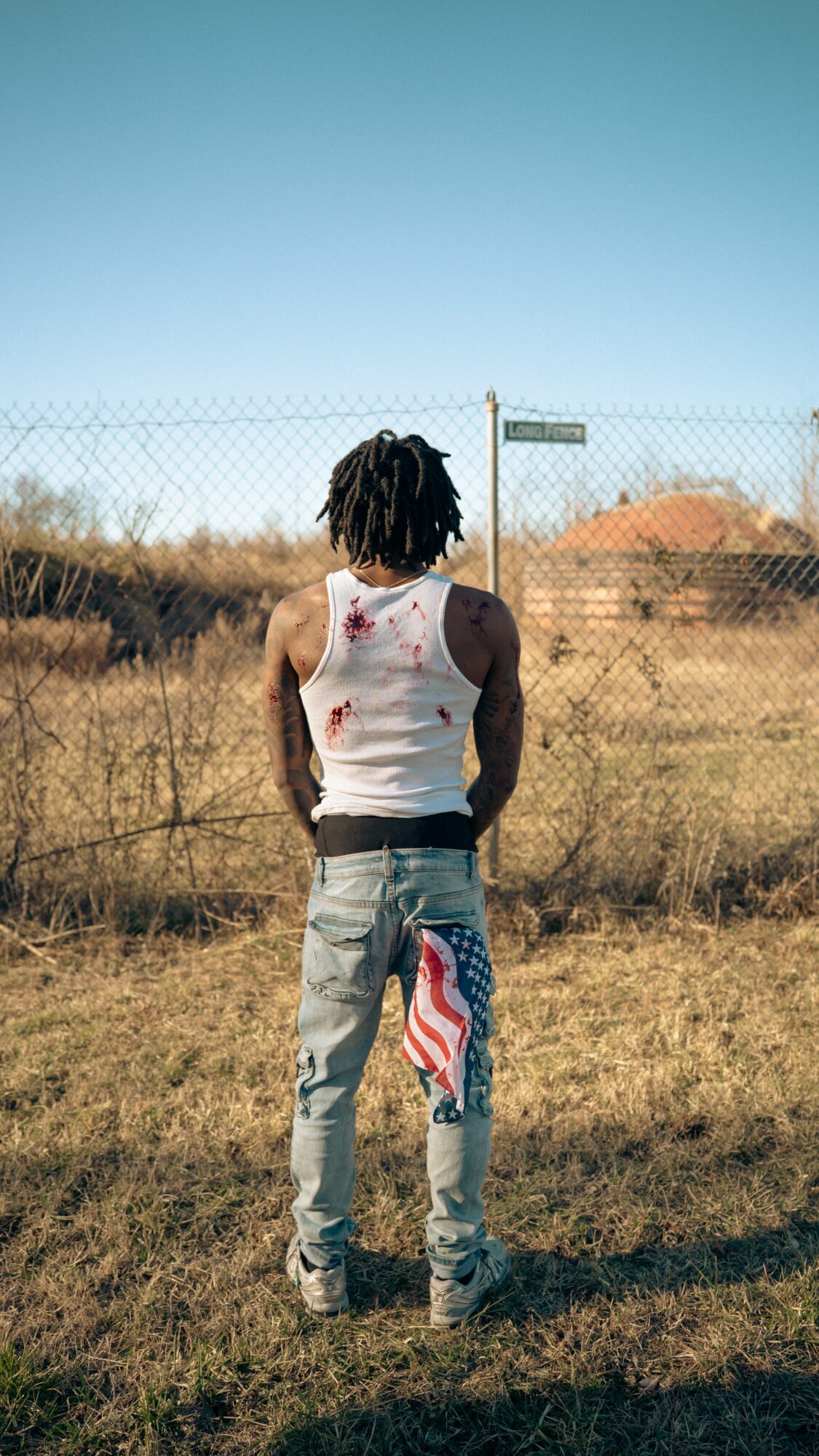 Person with dreadlocks wearing a white tank top and gray pants stands outdoors facing away, with a fence and open landscape behind.