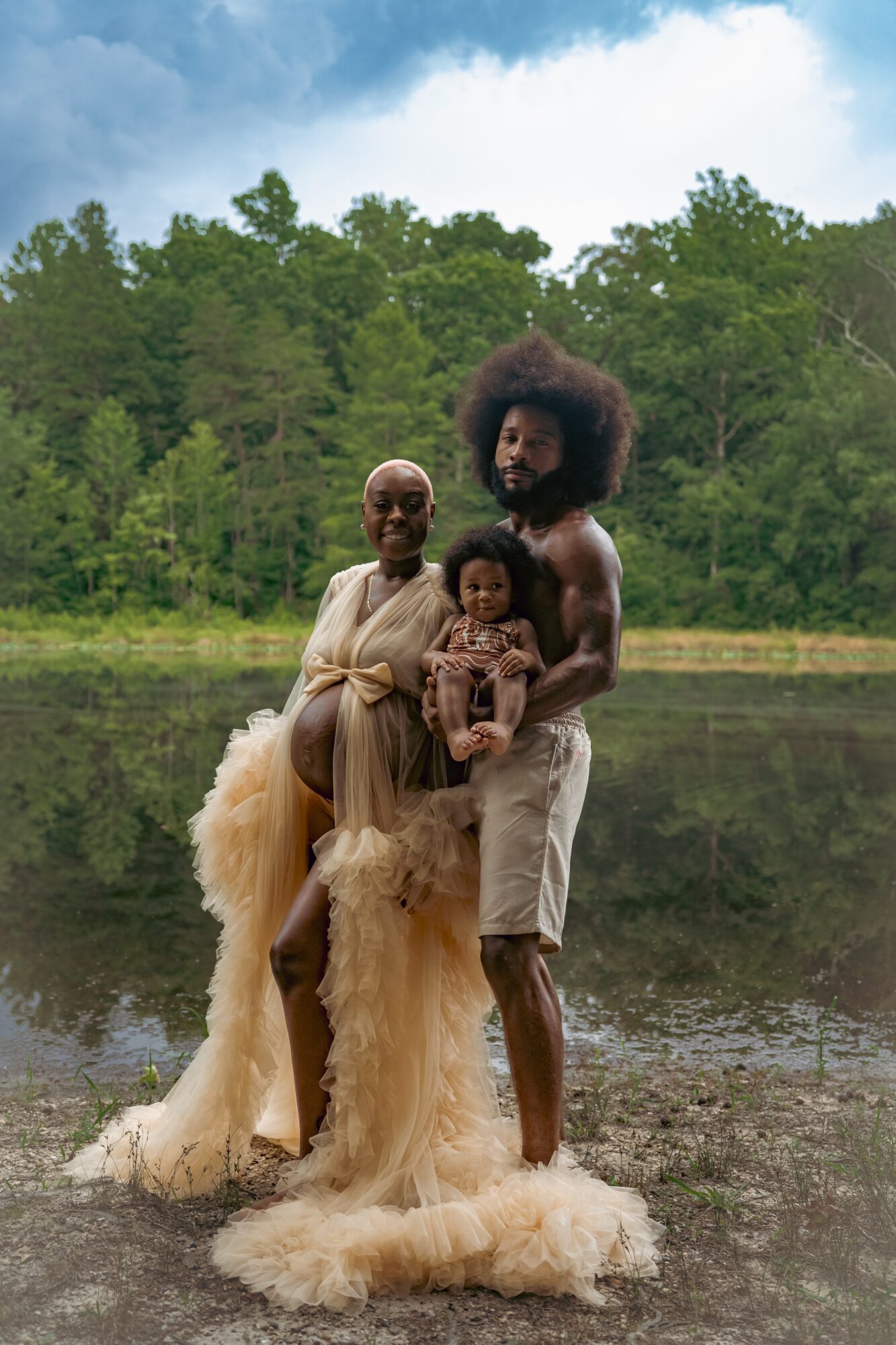 Three people standing by a lake with trees in the background, one holding a child, all smiling.