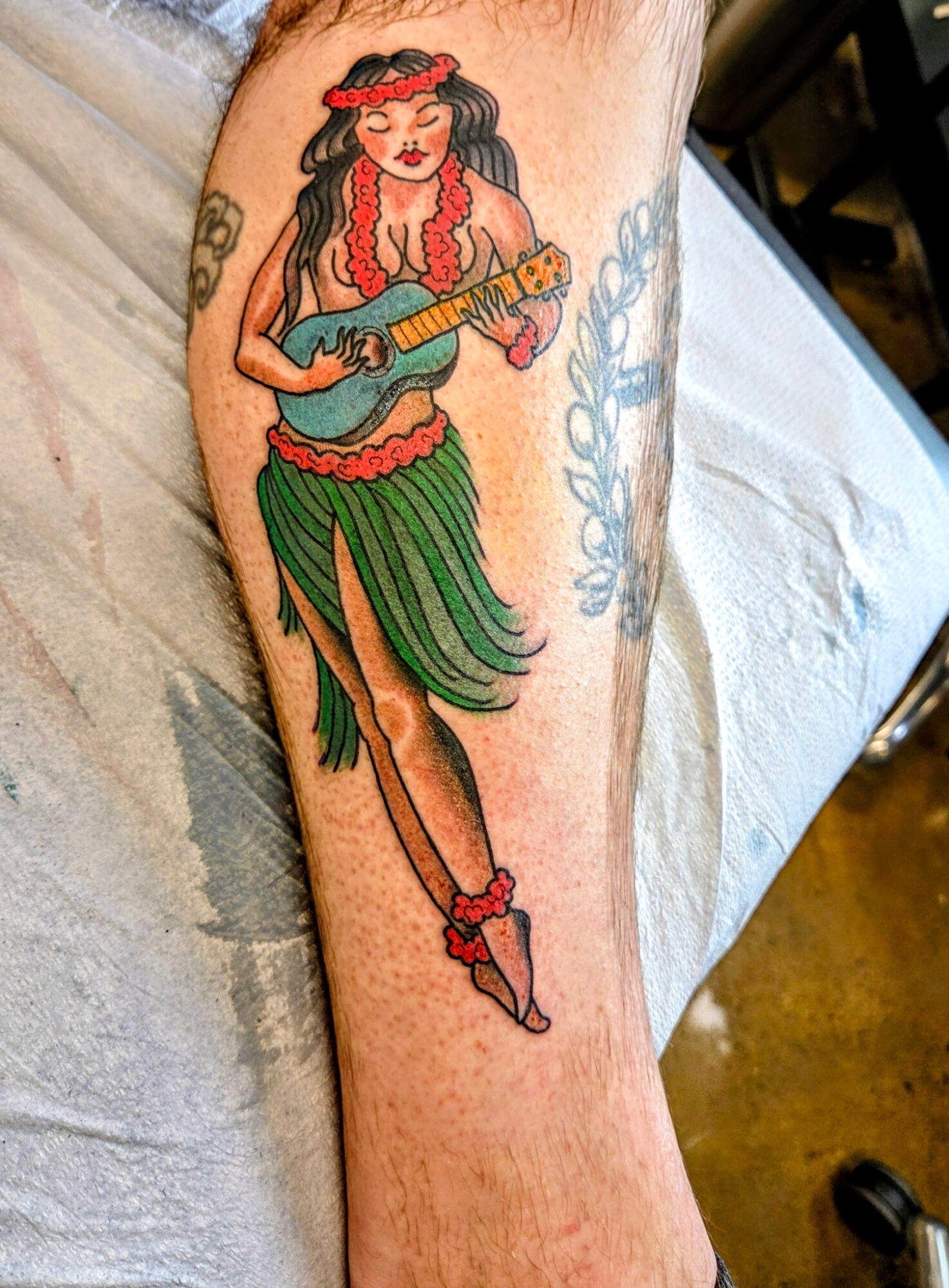 Tattoo of a woman in hula attire playing a ukulele, with a flower headband and grass skirt, on a person's arm.