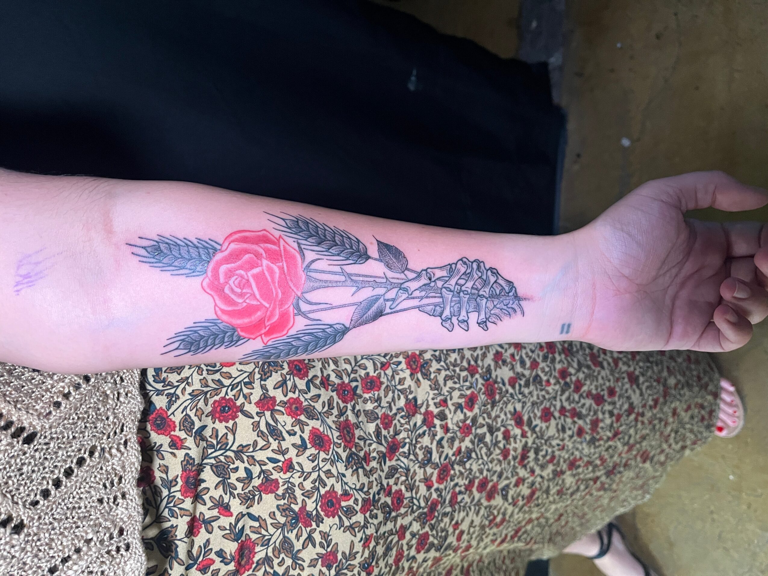Forearm tattoo of a skeleton holding a red rose with leaves, detailed and colorful, on a person's arm.