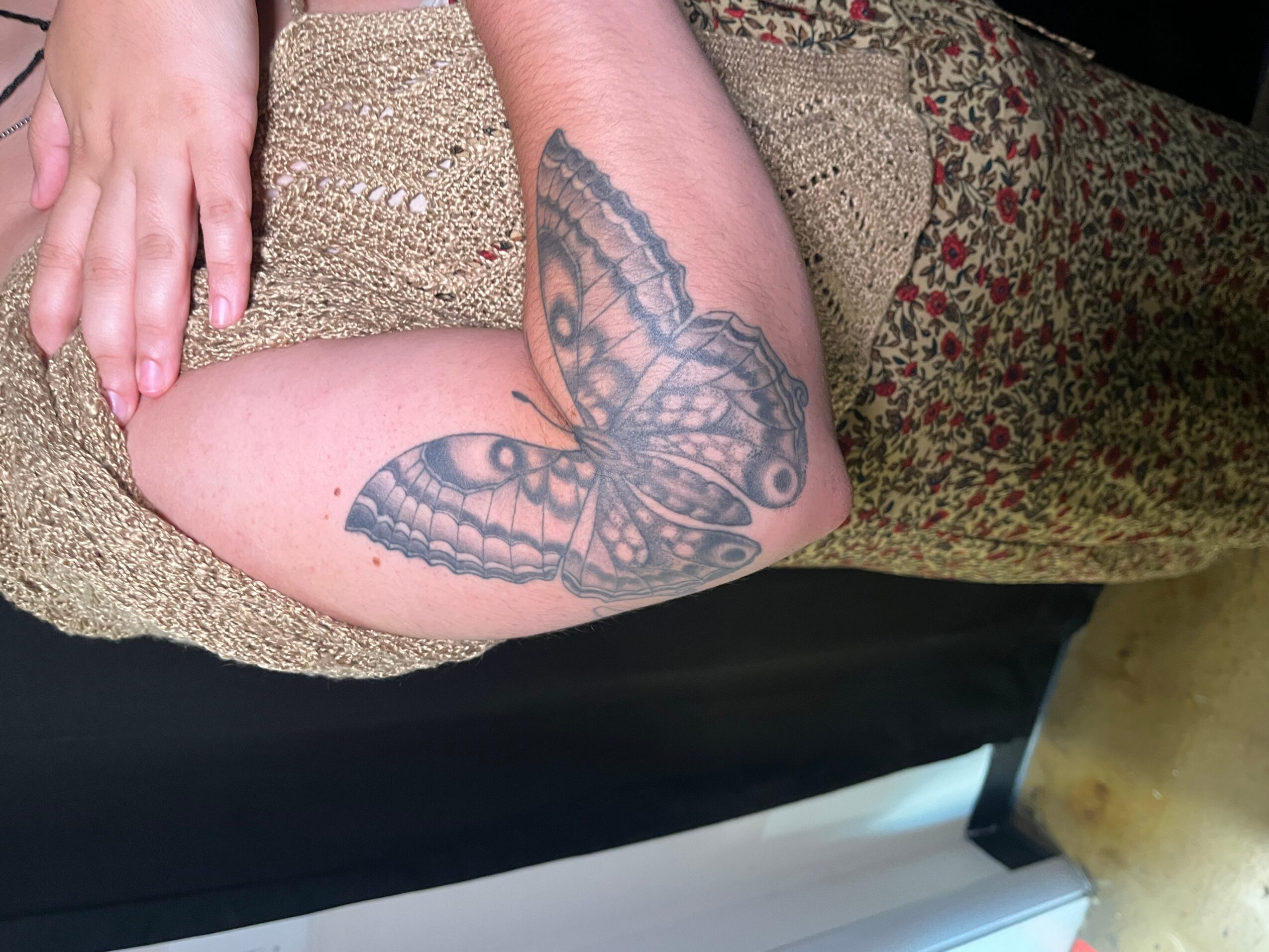 Person's arm with butterfly tattoo resting on textured fabric, with a black surface and a white object in the background.
